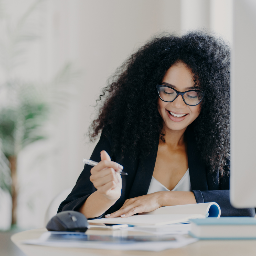 A woman with curly black hair and glasses smiling, seated at a desk with a computer, mouse, notebooks, and a pen.