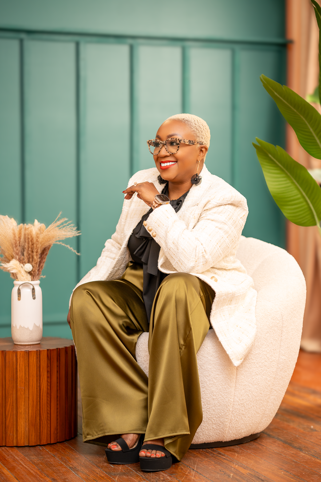 A woman with short blonde hair and glasses, wearing a cream blazer, black shirt, and shiny olive green pants, sitting on a cream-colored chair in a room with green walls and wooden flooring, smiling and showing a joyful expression.