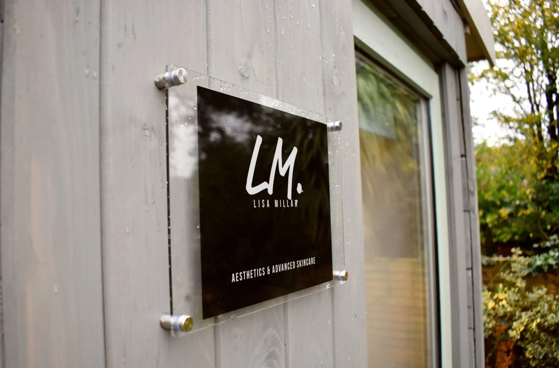 Exterior view of a skincare clinic with a black and white sign displaying the name 'LISA MILLAR' and the words 'AESTHETICS & ADVANCED SKINCARE' mounted on a wooden wall next to a window, with some water droplets on the sign and the wall.