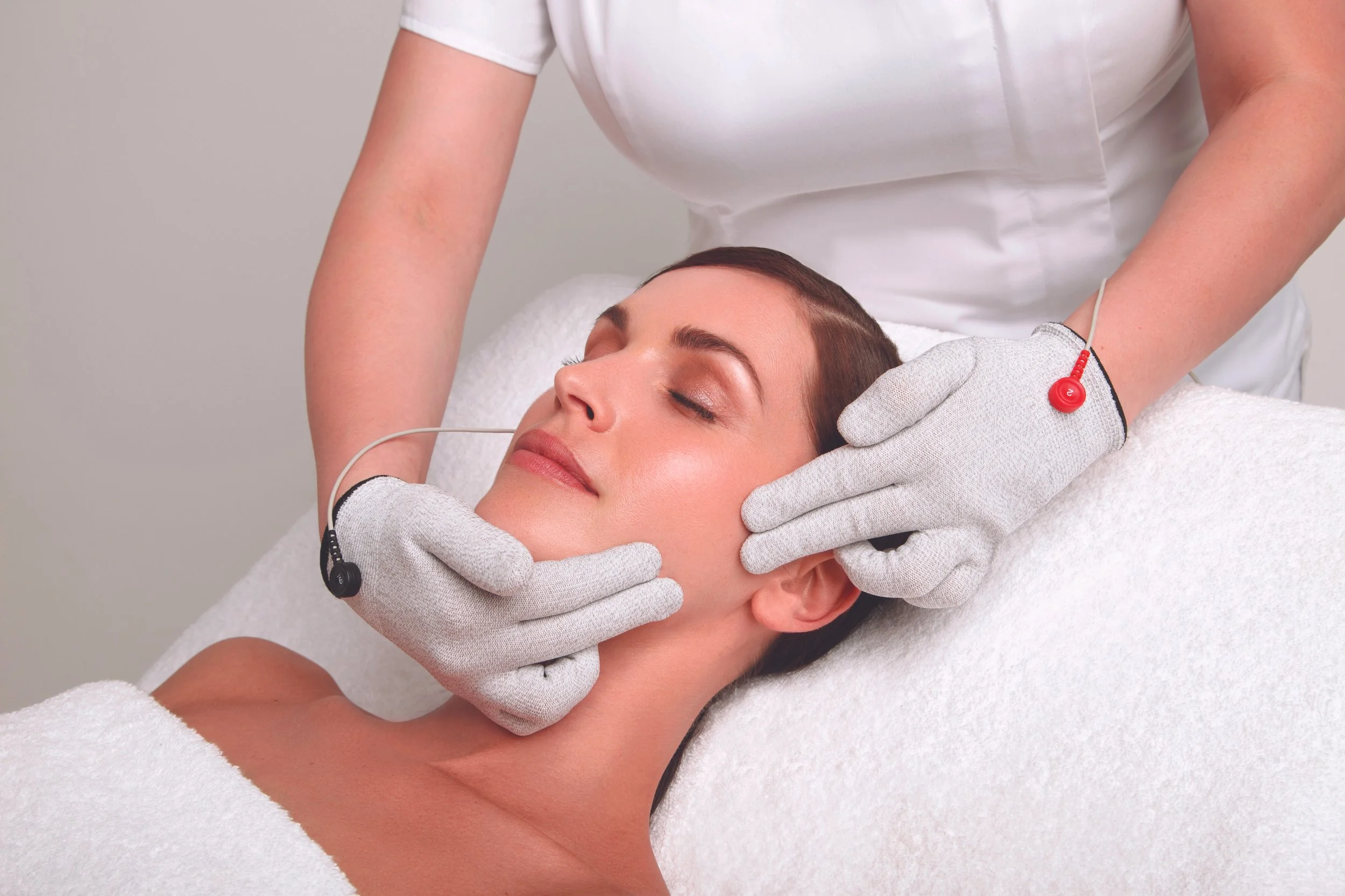 A woman receiving a facial treatment in a spa or clinic, with her eyes closed and a relaxed expression, while an aesthetician uses equipment on her face.