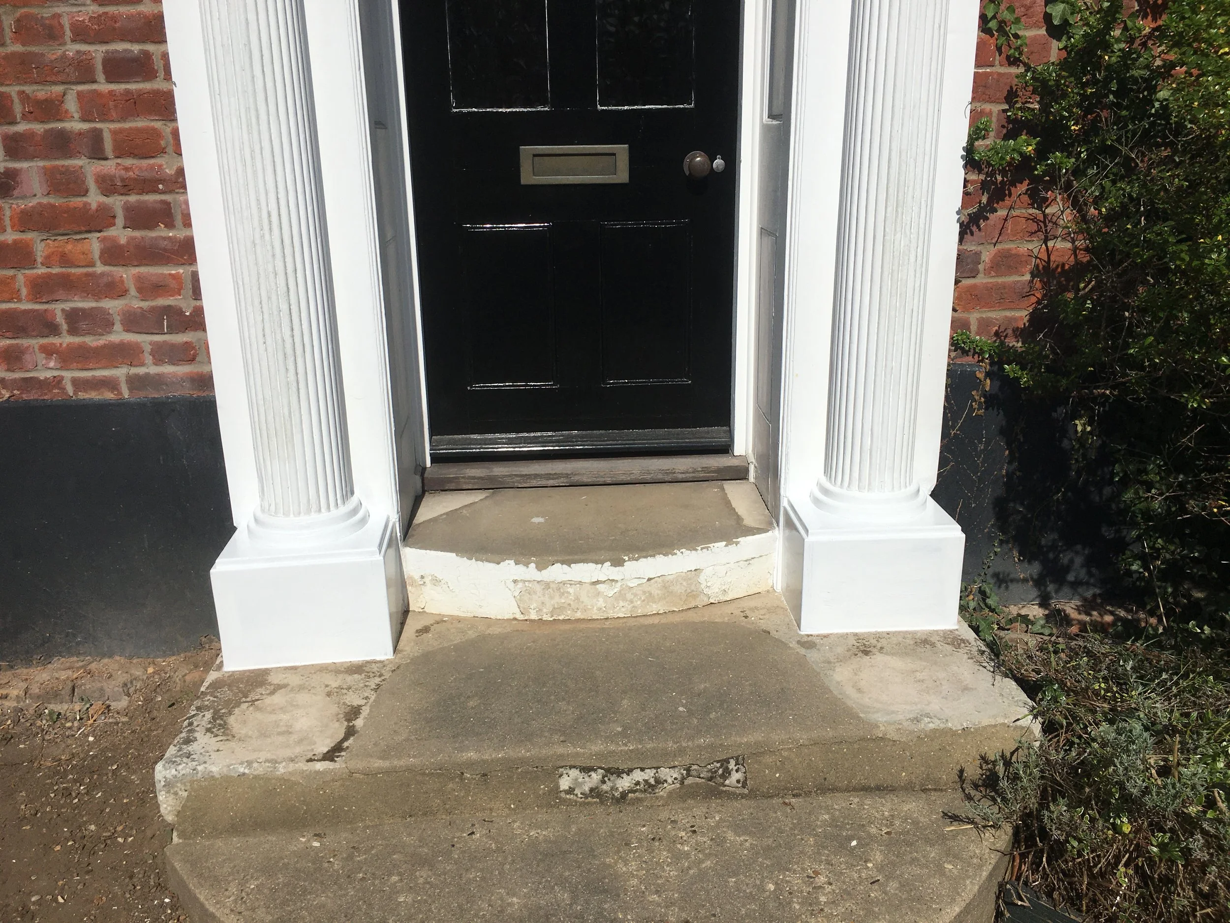 Front door with black paint, on a small porch with stone steps, flanked by white decorative columns, brick wall on the left, and bush on the right.