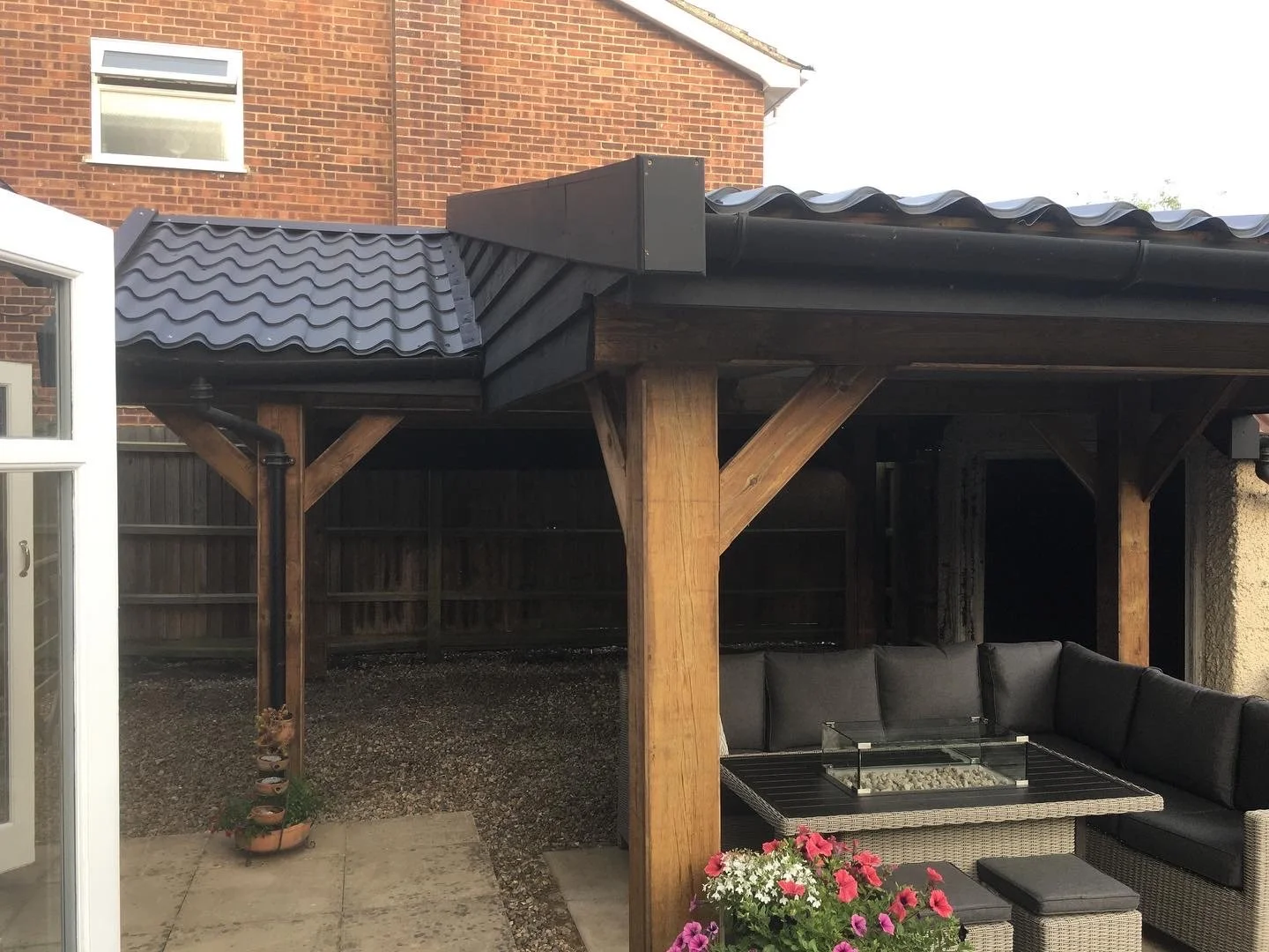 Backyard patio with a wooden pergola structure, granite fire pit table, black cushioned wicker sectional sofa, potted flowers, and a gravel ground area, with a brick house and a fenced yard in the background.
