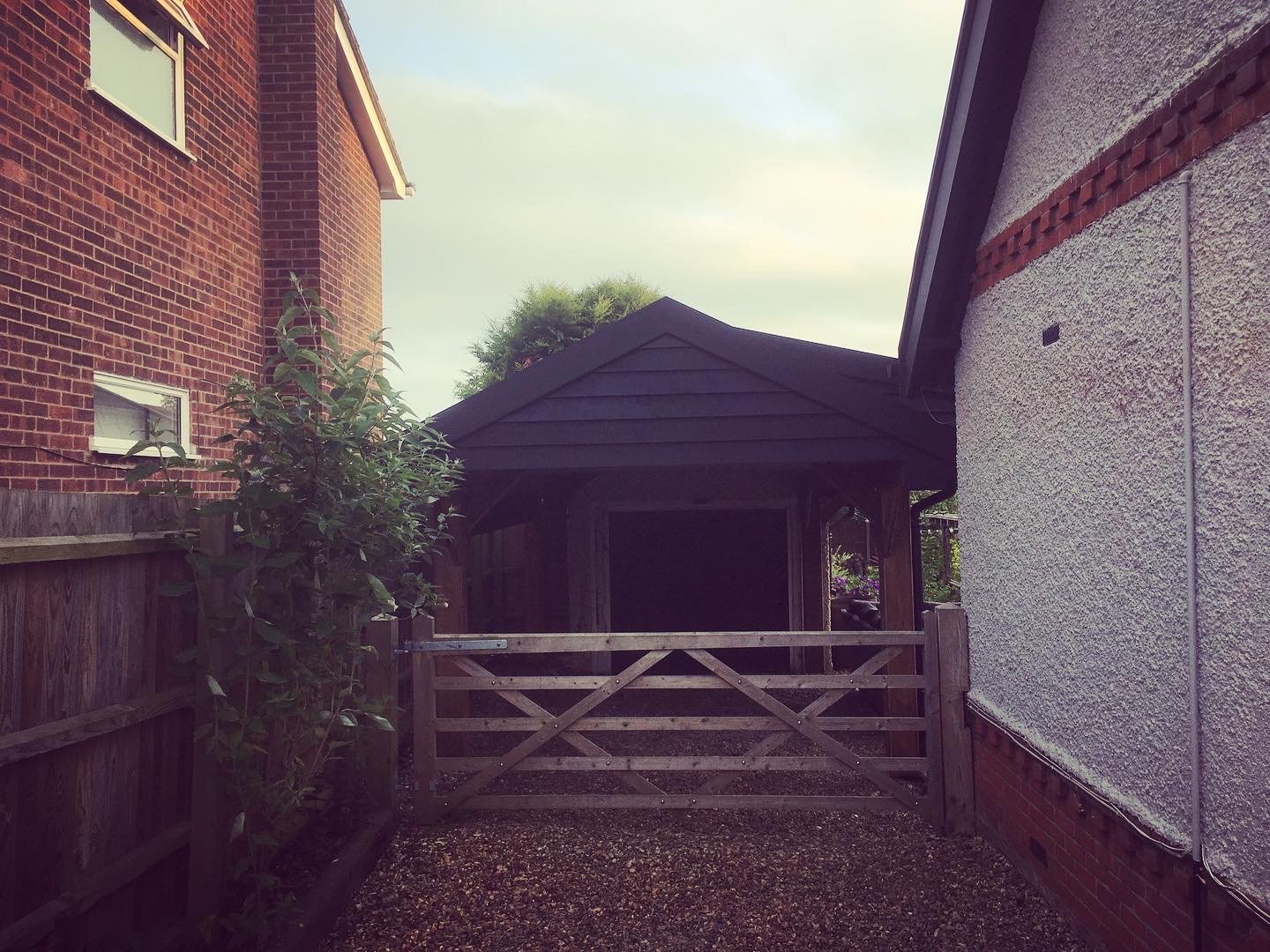 A backyard alleyway with a wooden gate, flanked by a brick house on the left and a stucco house on the right, with a small shed or gazebo in the background, under a cloudy sky.