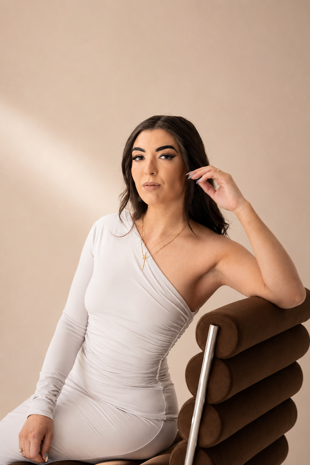 A woman with dark hair and makeup, wearing a white one-shoulder dress and gold jewelry, sitting on a brown modern chair against a beige background.