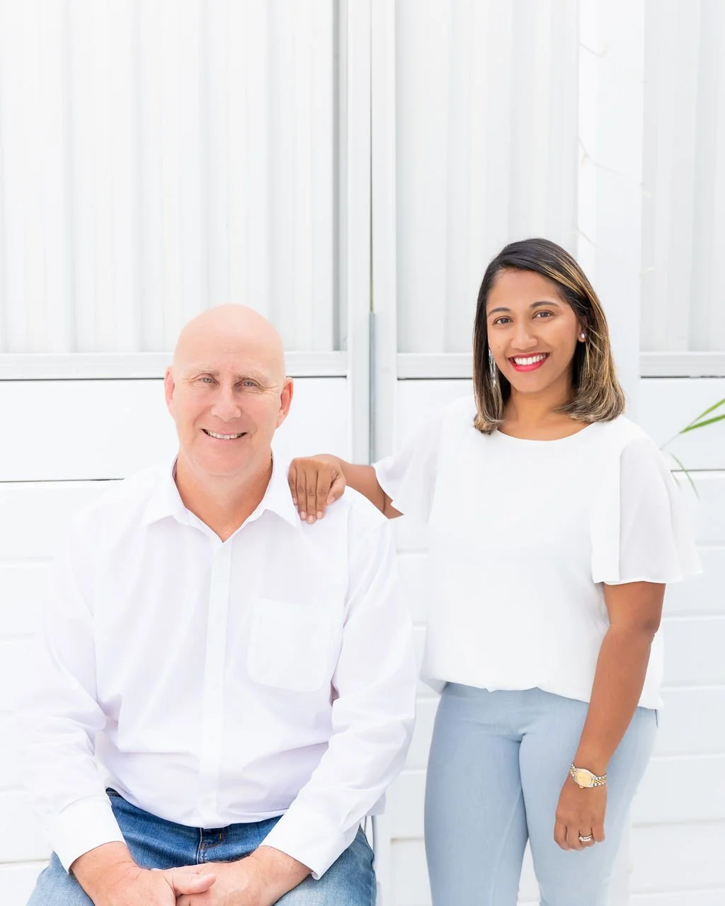 A man and woman smiling, posing indoors with a white background and a glass wall. The man is seated, wearing a white shirt and blue jeans. The woman is standing, resting her hand on the man's shoulder, wearing a white blouse and light blue pants, with jewelry and makeup.