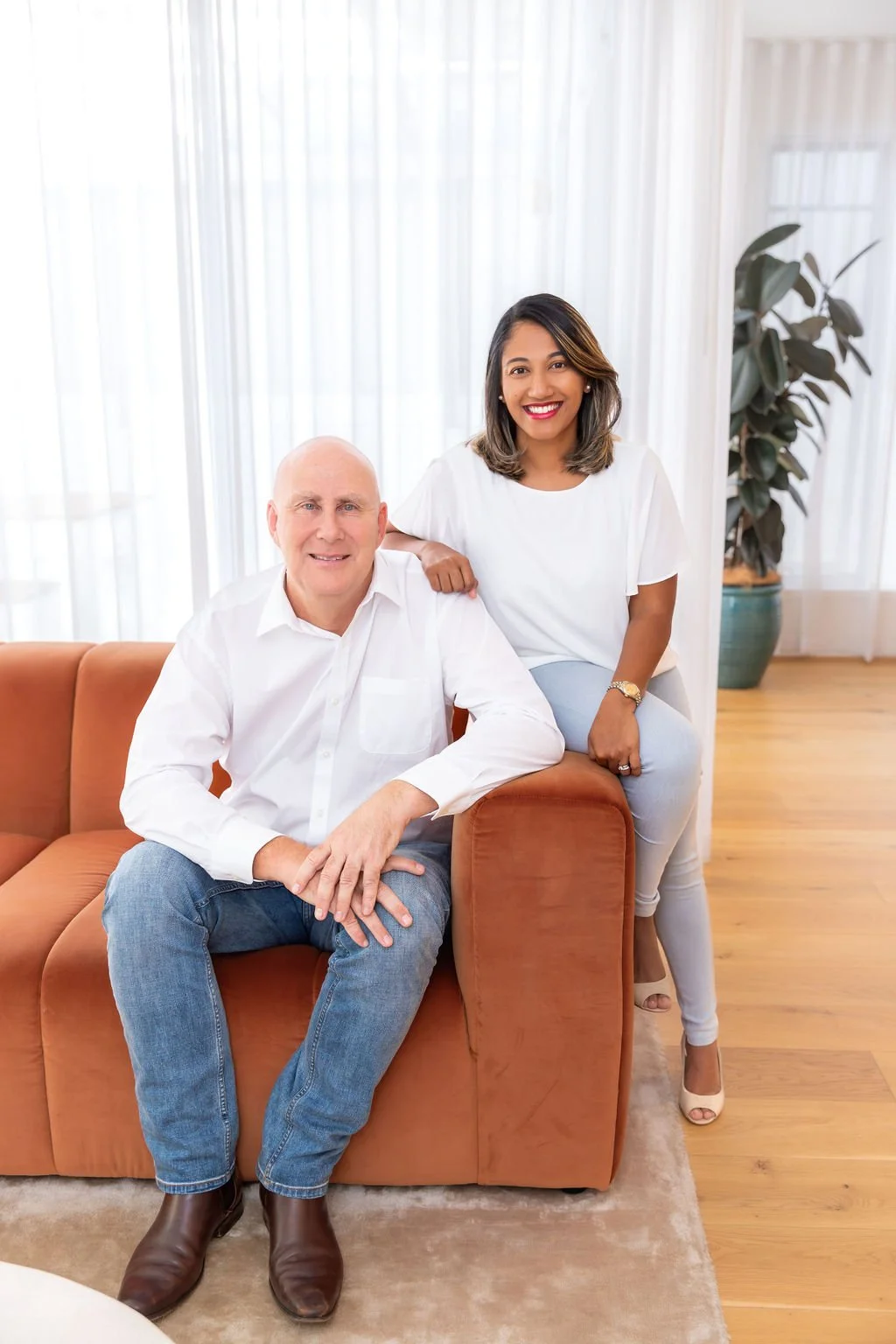 A man and a woman smiling and posing together in a bright room with white curtains and a large potted plant in the background. The man is seated on an orange couch, wearing a white shirt and blue jeans. The woman stands nearby with her hand on the man's shoulder, wearing a white shirt, light-colored pants, and high heels.