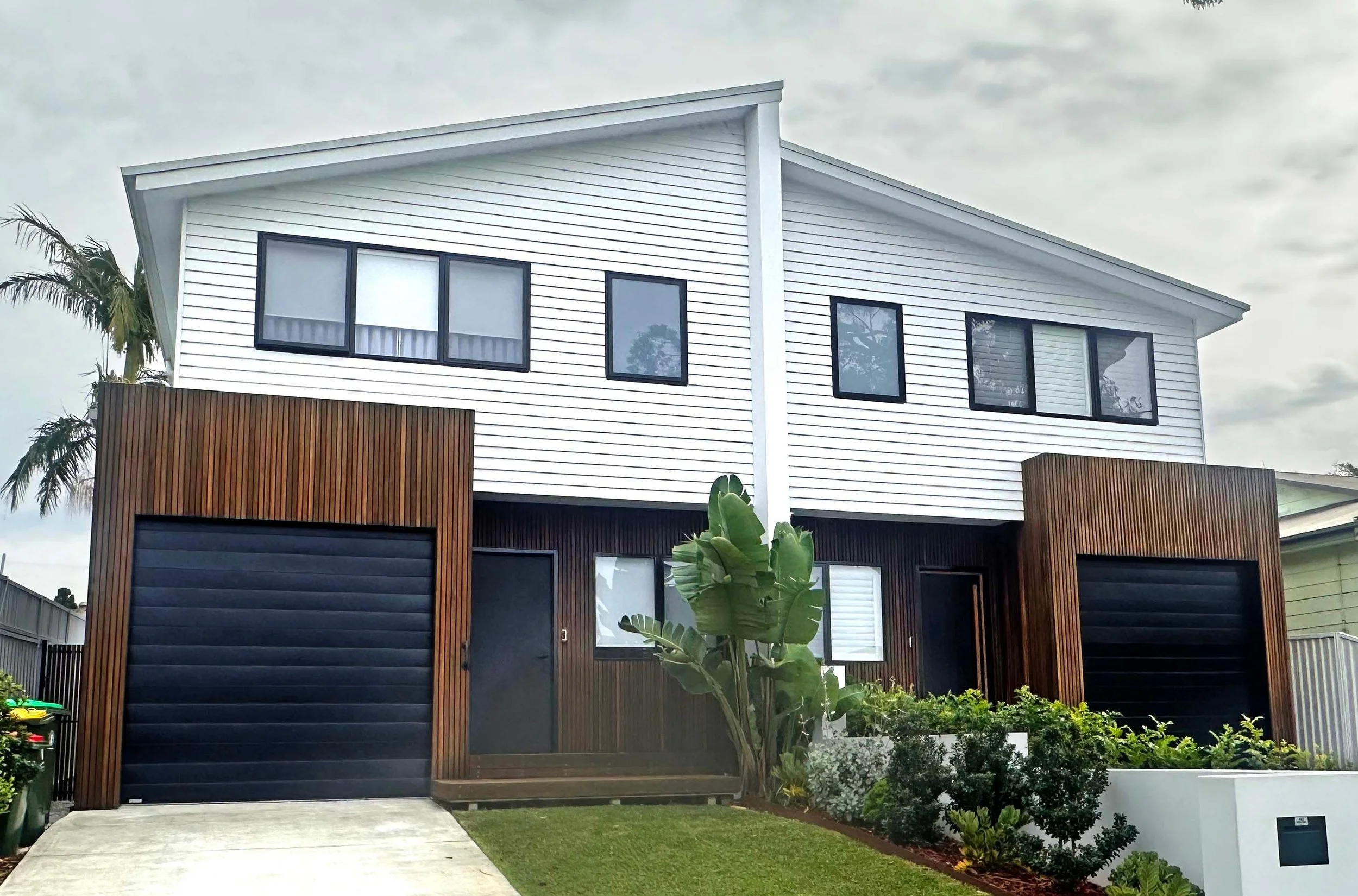Modern two-story house with a white upper exterior, black window frames, and brown wooden accents on the lower exterior, surrounded by greenery and palm trees.