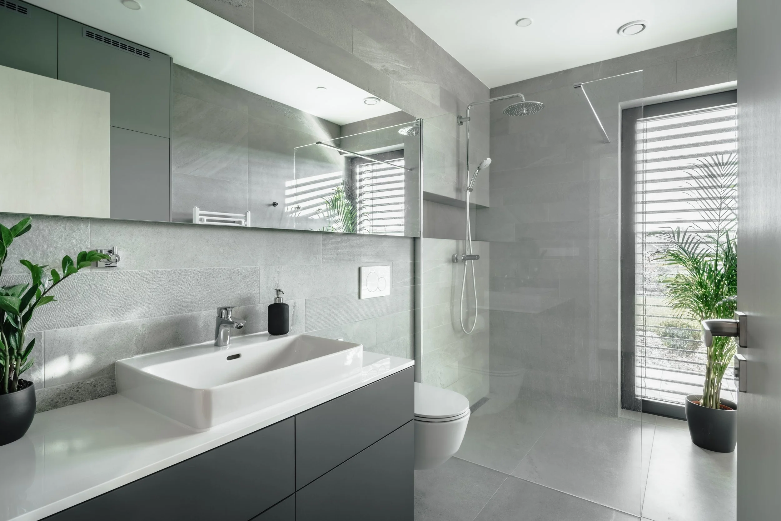 Modern bathroom with gray tiled walls and floor, white sink with black cabinet, toilet, large mirror, walk-in shower with rainfall and handheld showerheads, large window with blinds, and potted green plants.