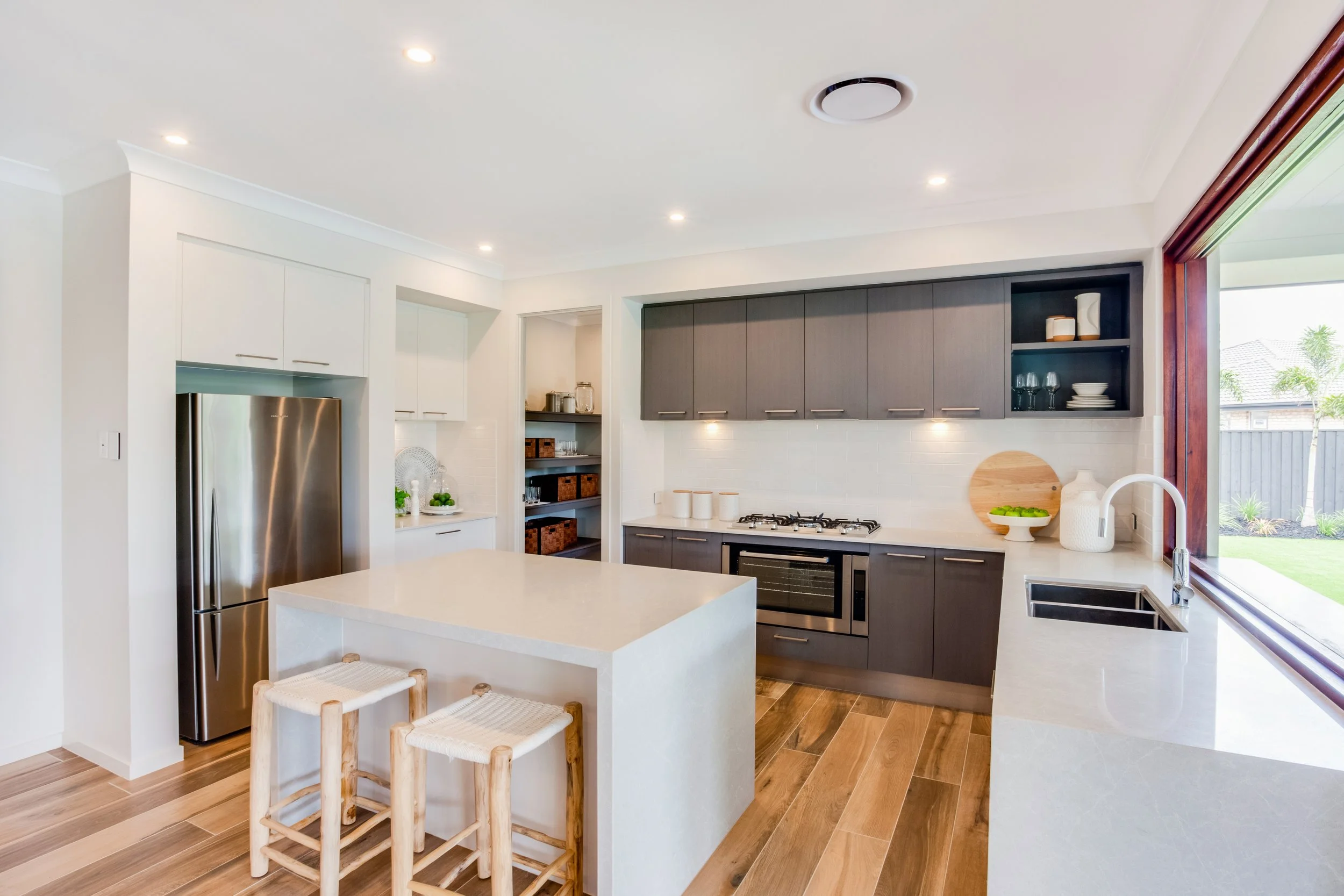 Modern kitchen with white and gray cabinetry, stainless steel refrigerator, island with two bar stools, built-in oven, gas stove, large window overlooking backyard, wooden floor, open shelving with dishes and glasses, minimal decor.