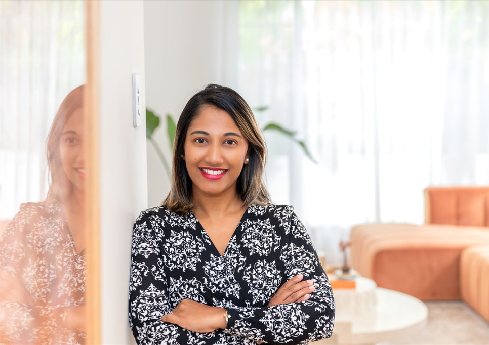 A woman with shoulder-length brown hair smiling with arms crossed, standing in a bright room with a couch and a potted plant in the background.