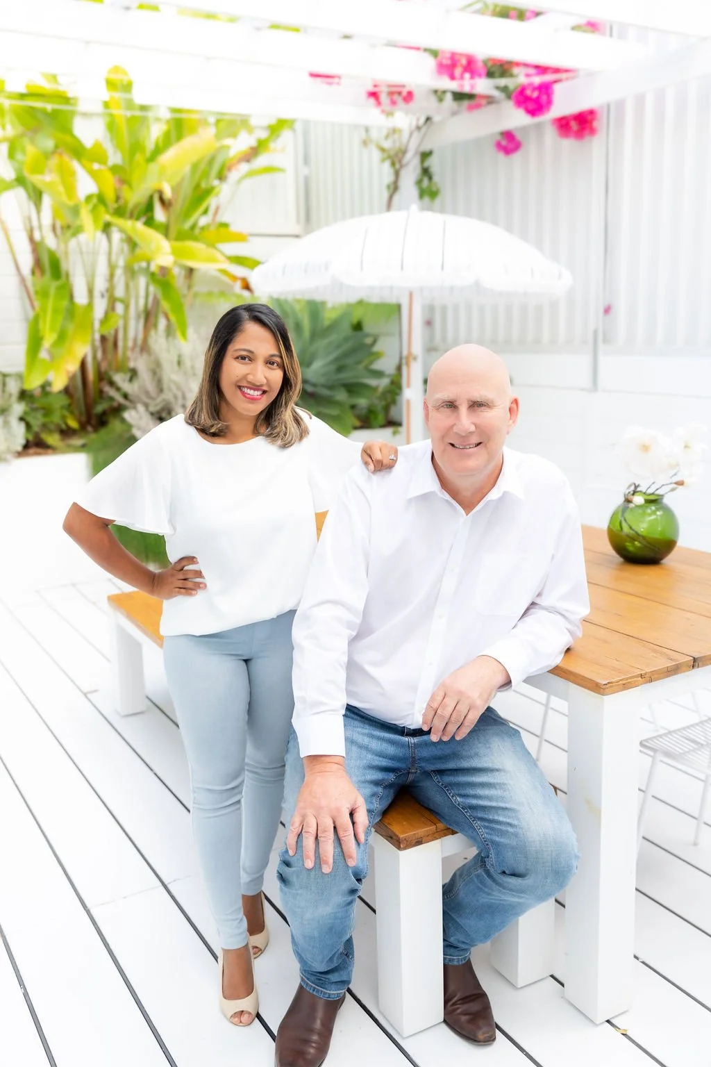 A smiling man and woman pose outdoors in a bright, white patio or garden area with green plants and pink flowers in the background. The woman stands with her hand on her hip, and the man is seated on a bench at a wooden table with a green vase and white flowers.
