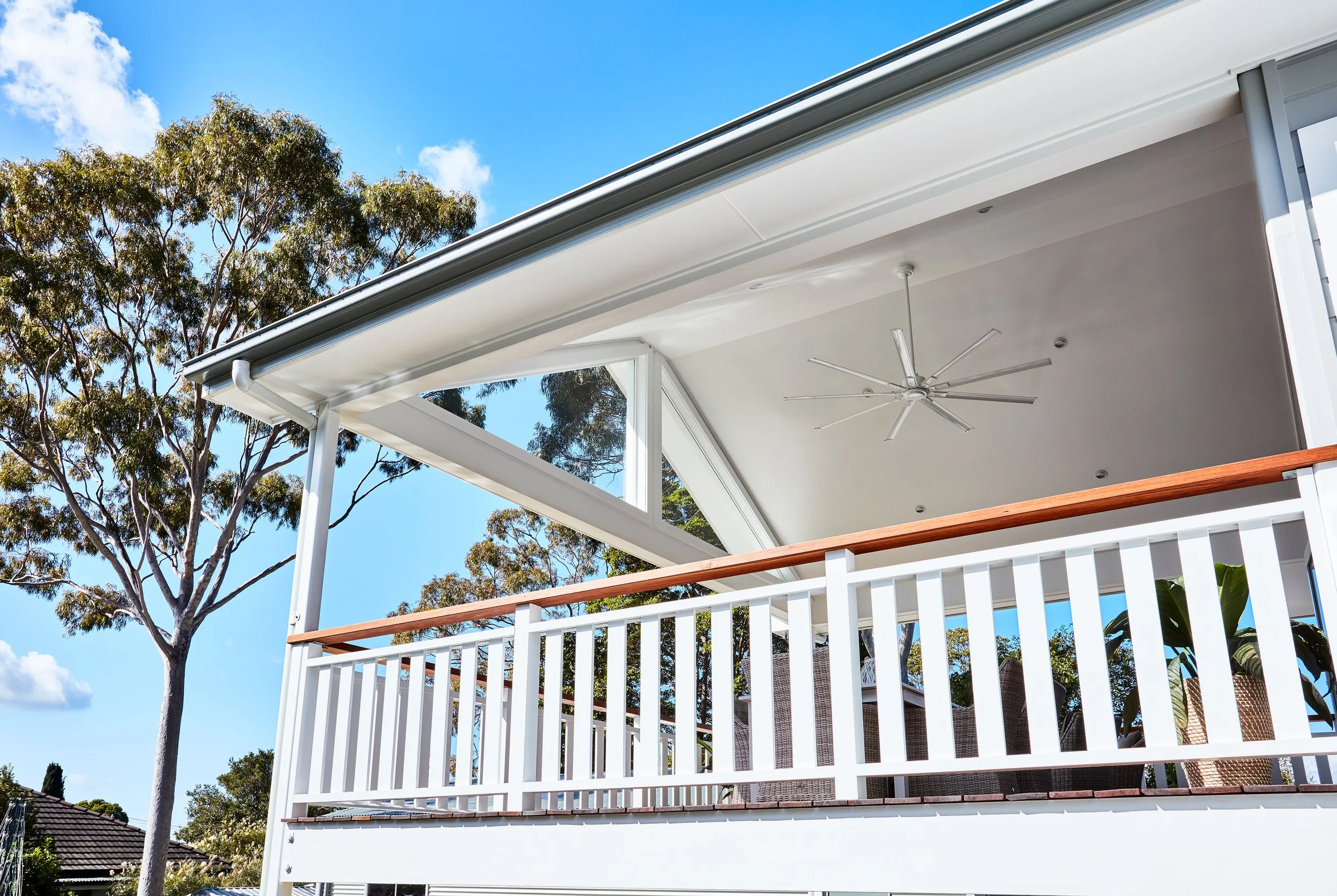 Upside-down view of a porch with a white railing, ceiling fan, and modern design, surrounded by trees against a blue sky.