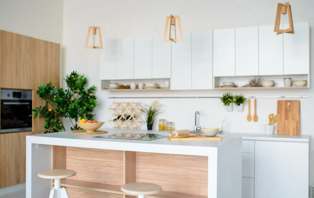 Modern kitchen with white cabinets, wooden accents, and an island with a white countertop. There are plants, bowls, hanging wooden utensils, and a cutting board. The lighting fixtures are wooden with simple geometric designs.