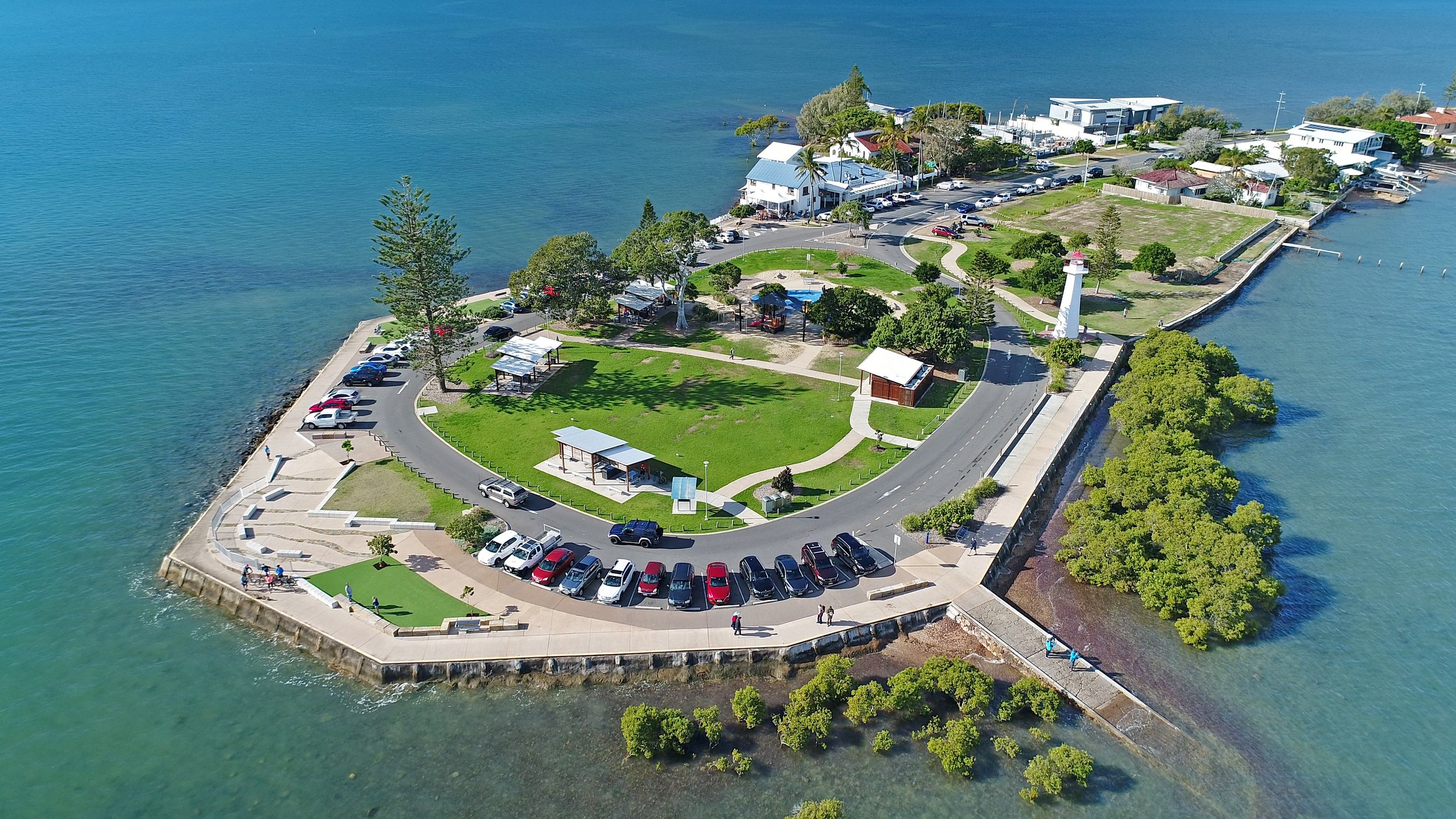 Aerial view of a small park with a lighthouse on a waterfront peninsula surrounded by water, with parking lots, walking paths, trees, small structures, and buildings nearby.