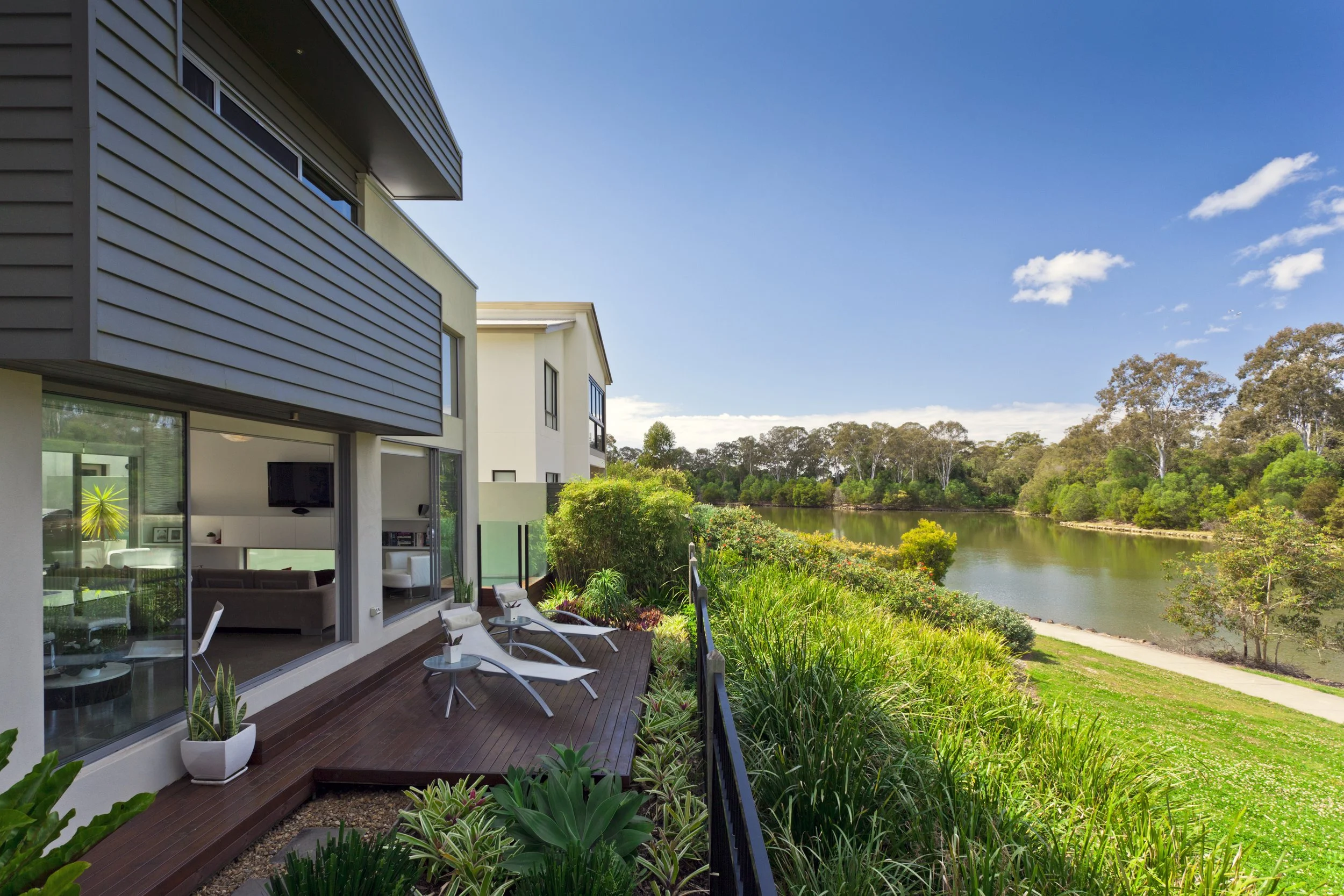 Modern apartment building with a balcony overlooking a river and lush greenery on a sunny day.