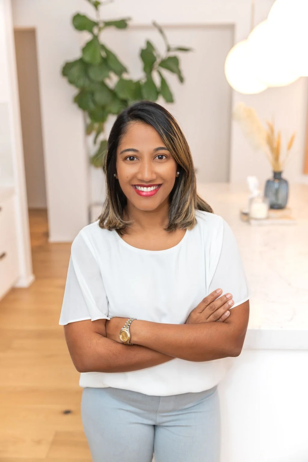 A woman with shoulder-length hair, smiling, standing with arms crossed in a bright room with a plant and decorative items in the background.