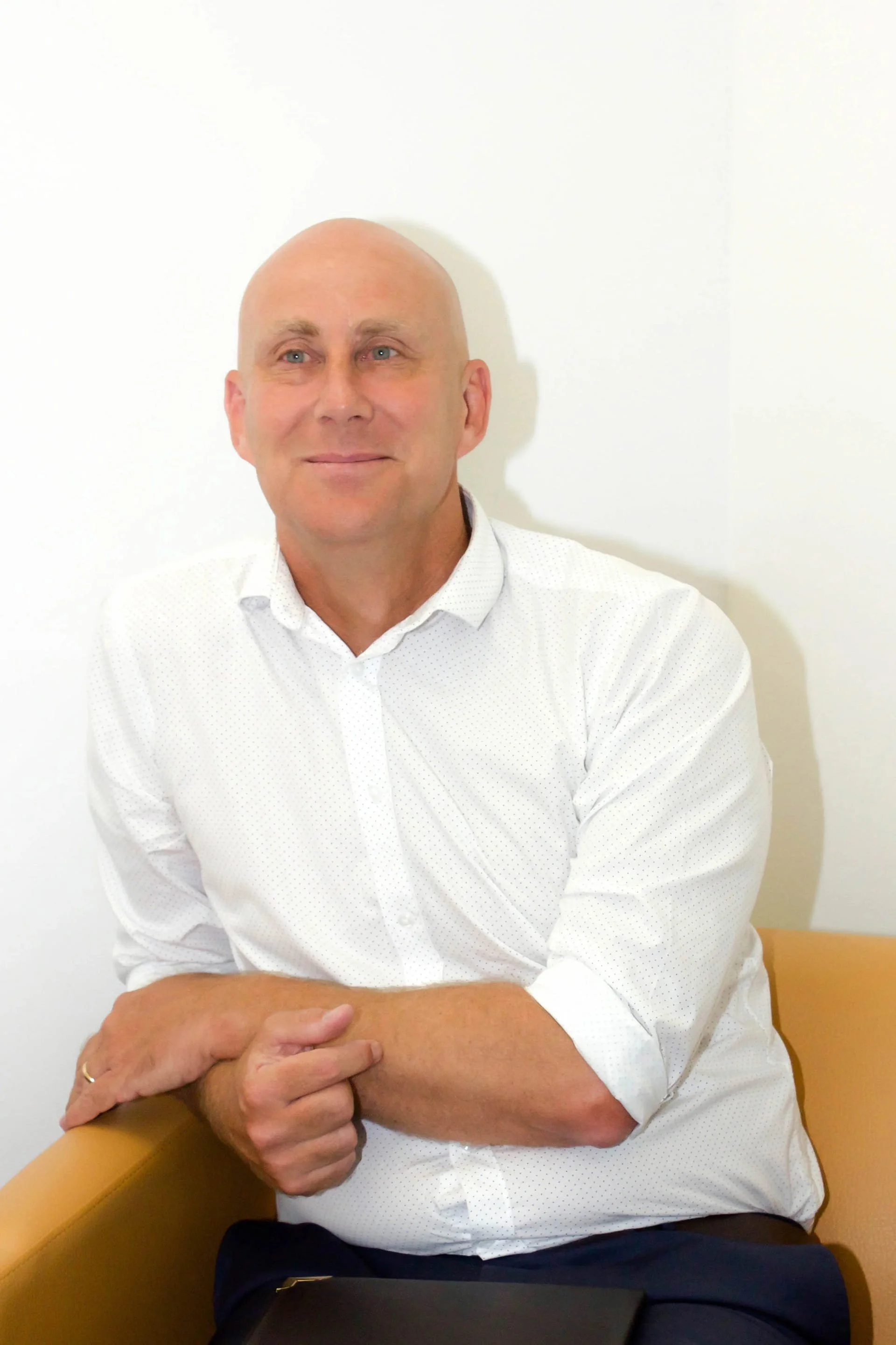 A bald man with light skin, wearing a white dress shirt with small dots, sitting on a tan chair against a white wall, looking slightly to the left with a slight smile.