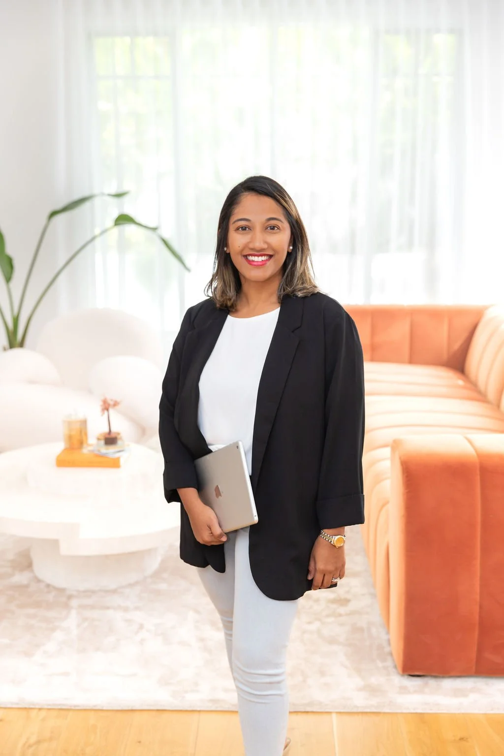 A woman in a black blazer and white top holding a silver laptop standing in a bright, modern living room with a peach-colored sofa, a white coffee table, and a large window with sheer curtains.
