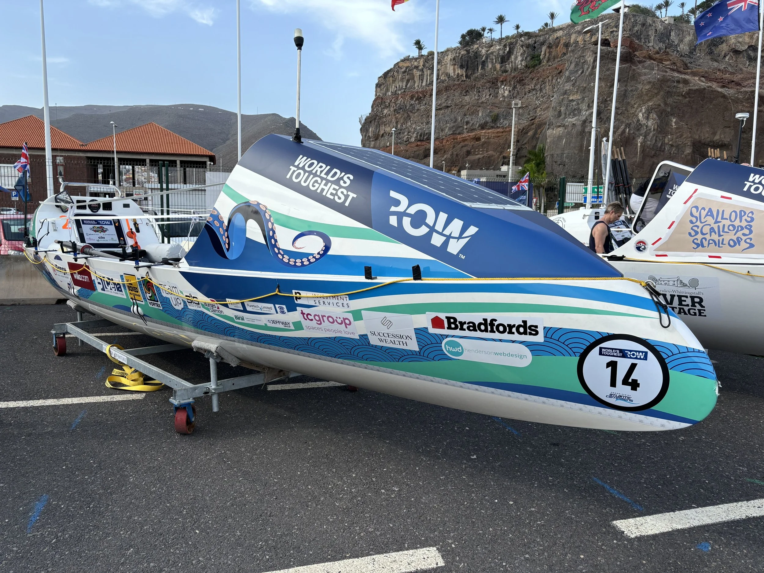 A speedboat with sponsor logos and the number 14, displayed on a wheeled platform in an outdoor parking lot with other boats and flags in the background.