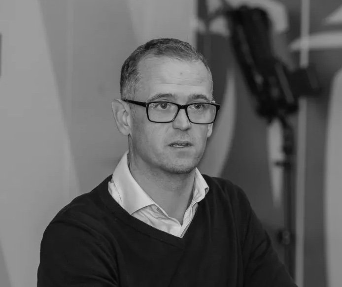 A man with glasses wearing a collared shirt and sweater, sitting indoors with a serious expression.