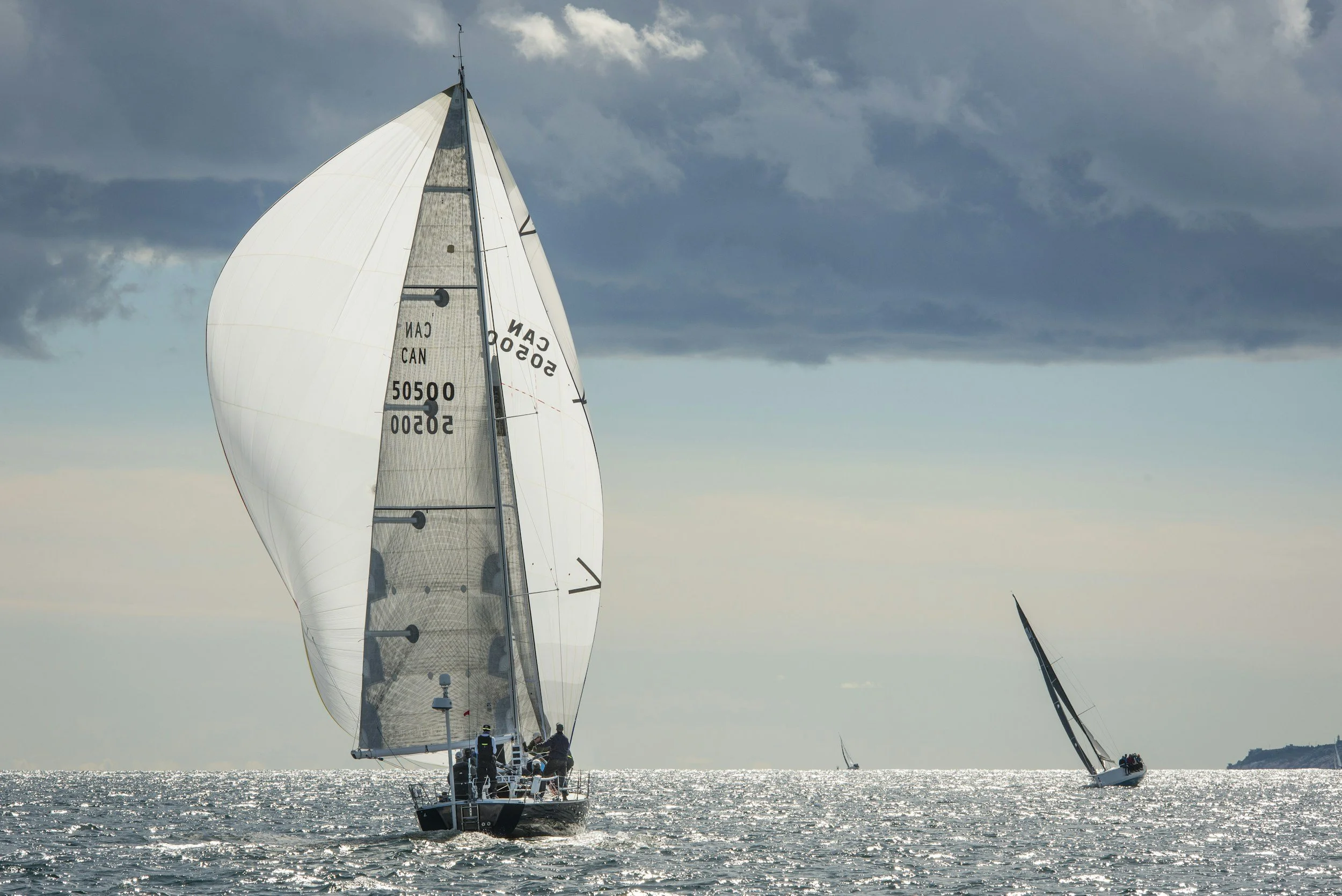 A sailboat with a large white sail glides across the shimmering water under a cloudy sky, with two other sailboats visible in the distance.