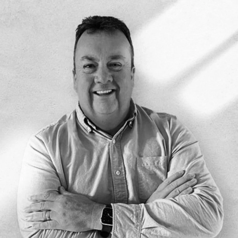 Black and white photo of a smiling man with crossed arms, wearing a collared shirt and a wristwatch, standing against a plain wall.