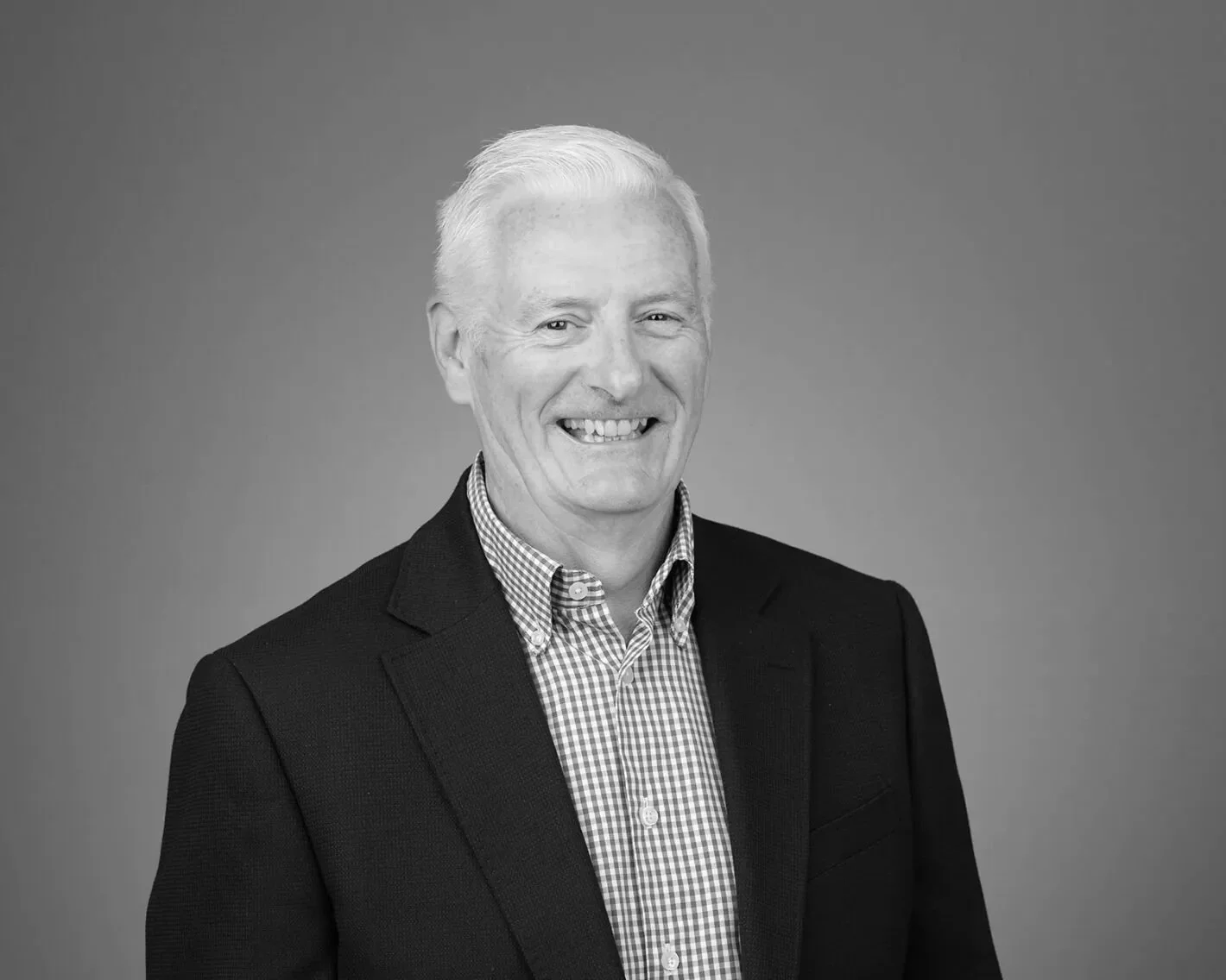 Black and white portrait of a smiling middle-aged man with short light hair, wearing a dark blazer and checkered button-up shirt against a plain background.