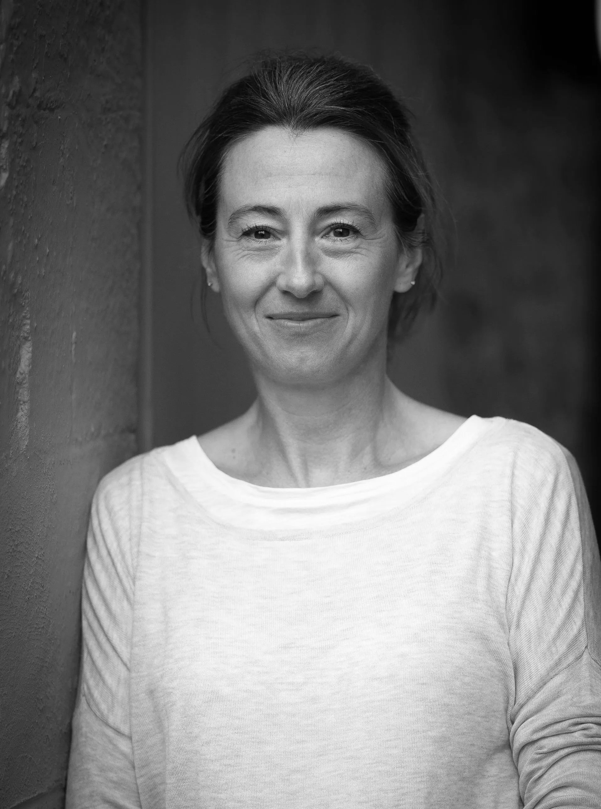 Black and white portrait of a woman standing outdoors, leaning against a brick wall, smiling softly, wearing a light-colored long-sleeve shirt.