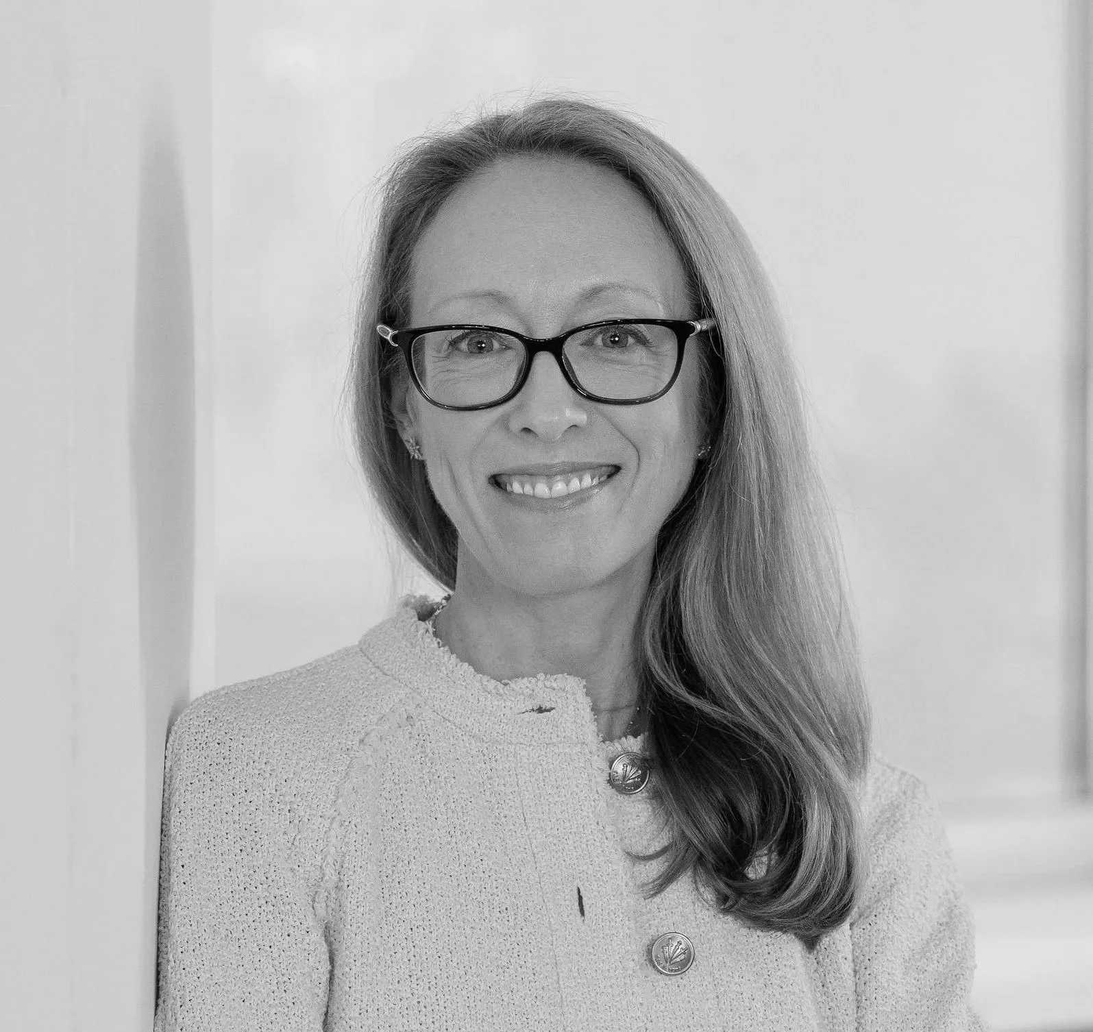 Black and white portrait of a woman with long hair, glasses, smiling, wearing a textured light-colored blazer with decorative buttons, standing indoors.