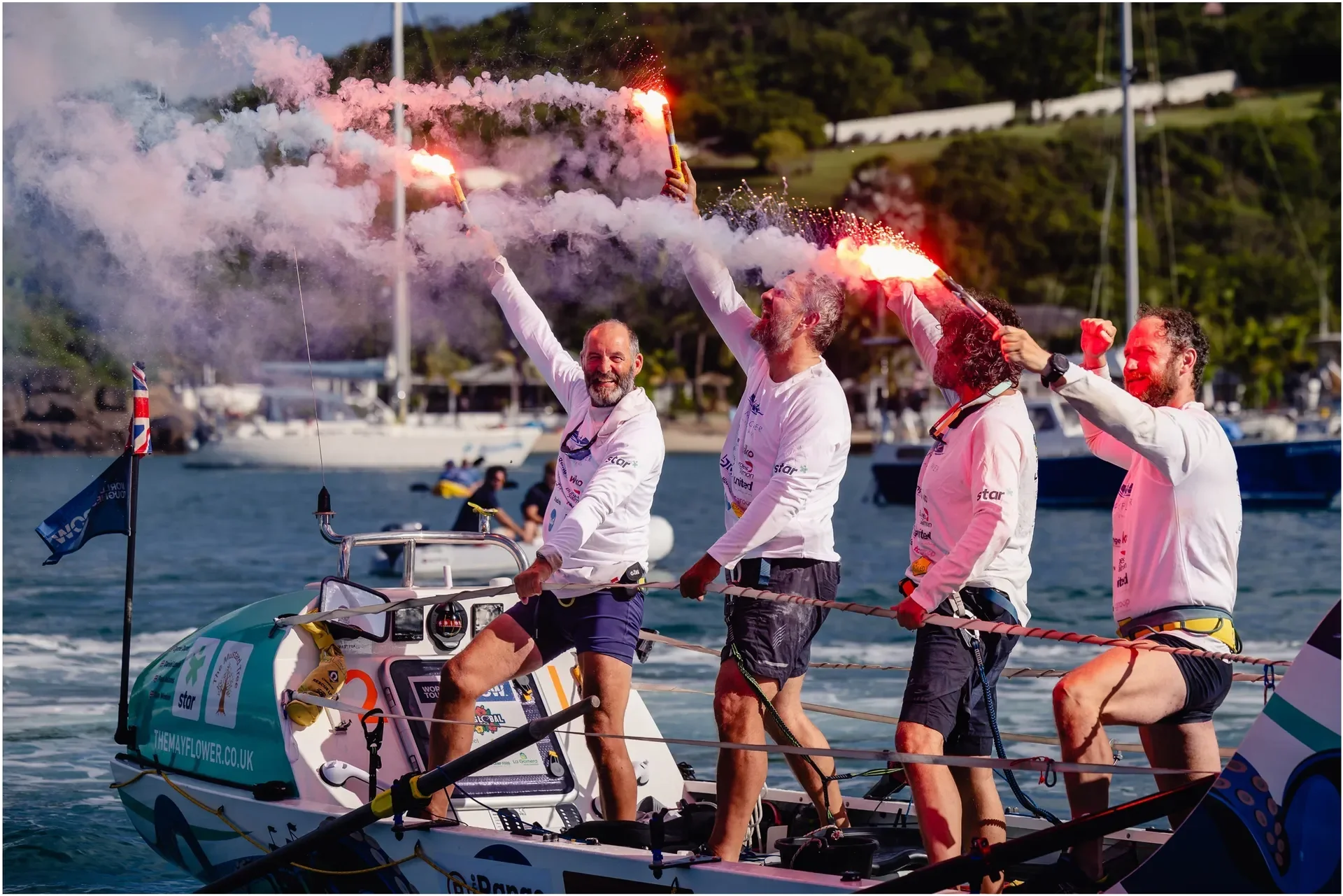Four male sailors on a boat celebrating victory with smoke flares in their hands, smiling and cheering, during a sailing race near a marina with boats and greenery in the background.