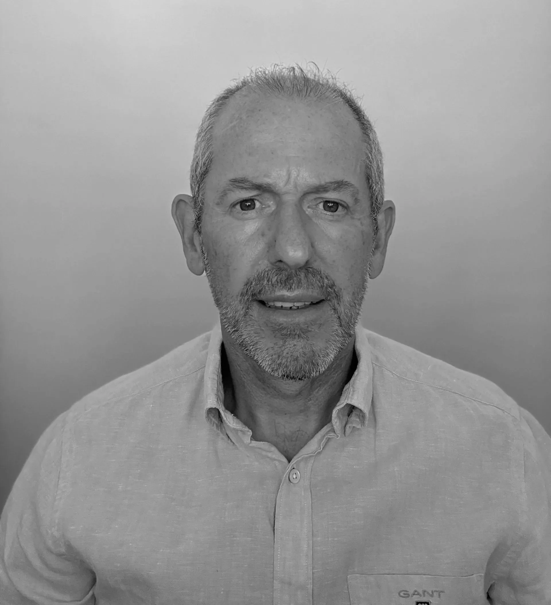 A black and white photo of a middle-aged man with short, gray hair, a beard, and mustache, wearing a light-colored collared shirt.