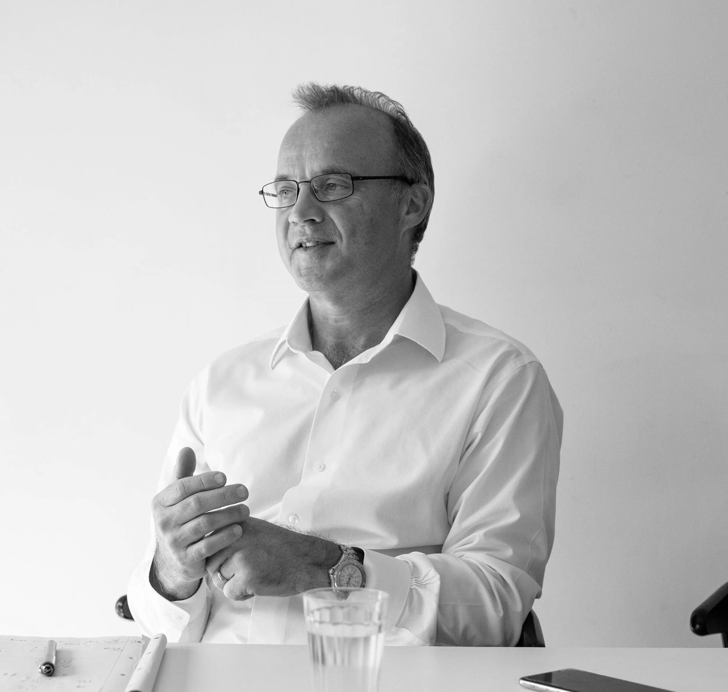 Black and white photo of a man with glasses in a white dress shirt sitting at a table with a glass of water, a pen, and a smartphone.