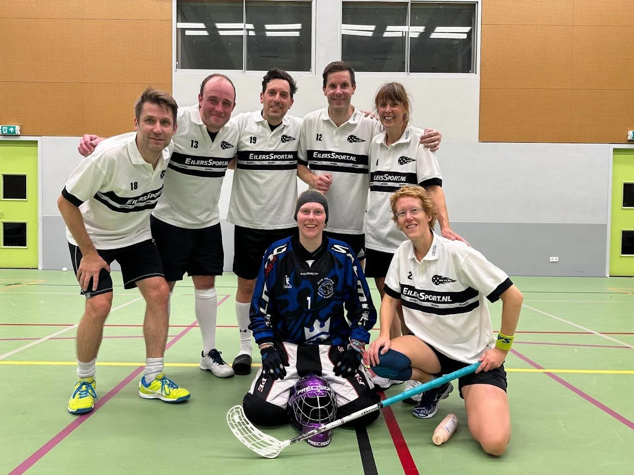 A group of seven floorball players, six standing and one kneeling in front, on an indoor court. They are wearing team jerseys, and the kneeling player is a goalkeeper with gloves and protective gear. The team is smiling and posing for a team photo.
