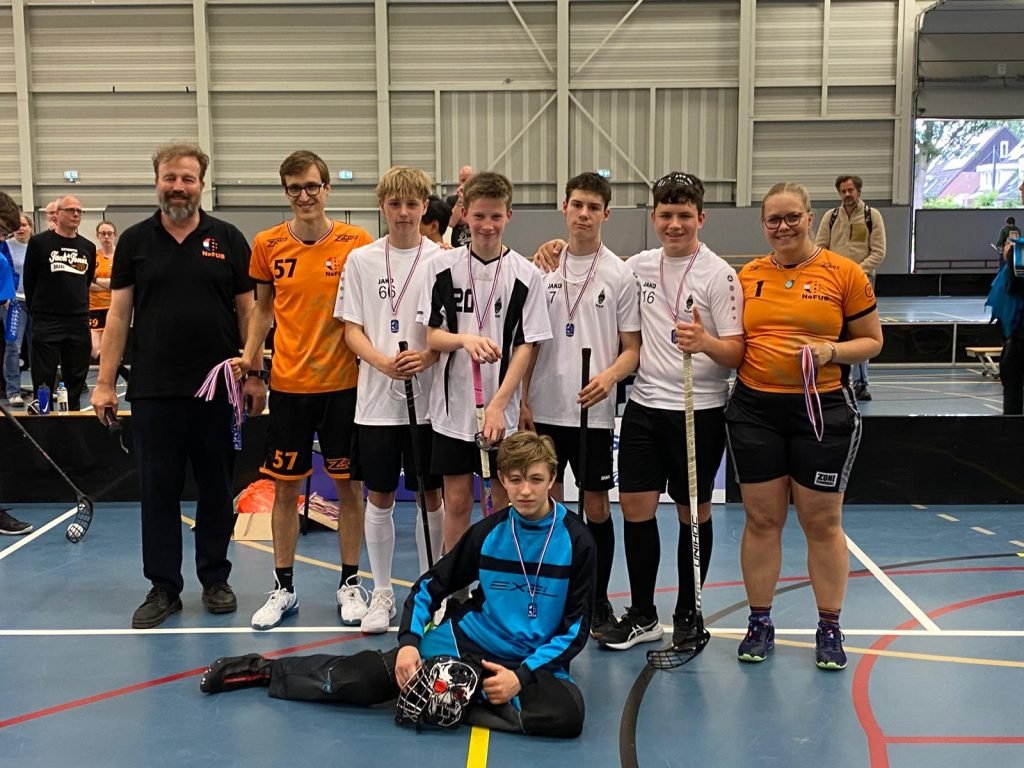 A group of young male floorball players and two coaches posing together in an indoor sports hall, some holding medals and floorball sticks, celebrating after a game or tournament.