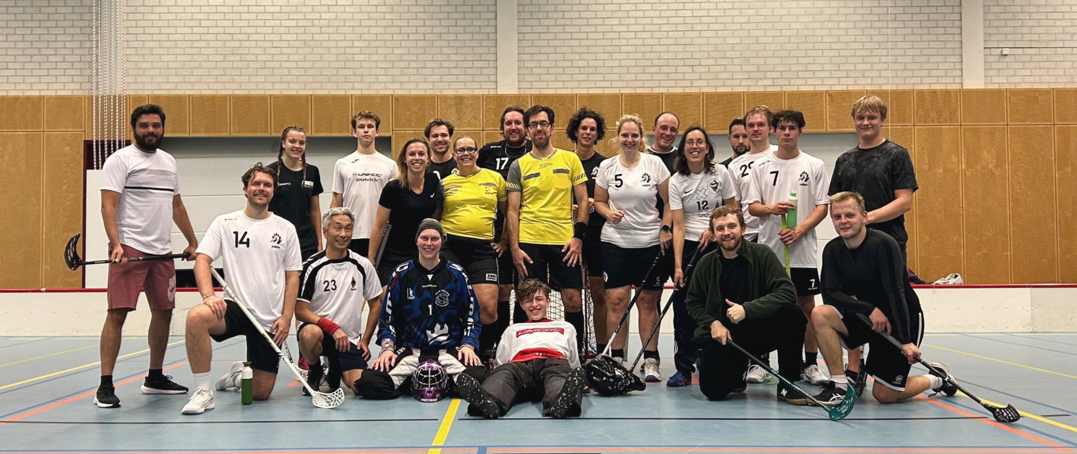 A group of people posing for a team photo on an indoor floor hockey rink, some holding floor hockey sticks and wearing sports jerseys.