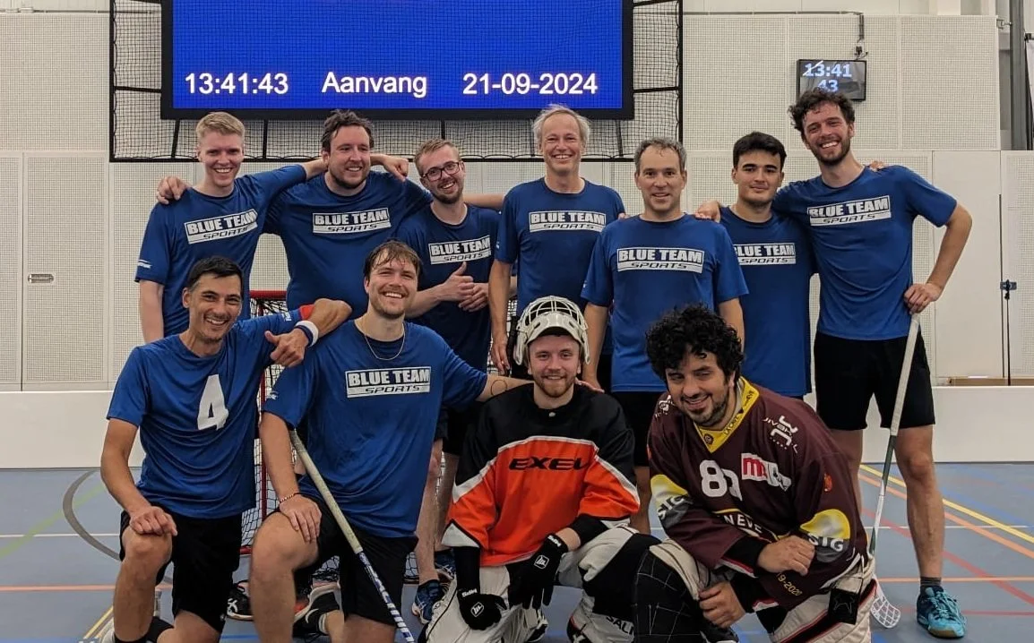 A group of ten men in sports uniforms posing together in an indoor sports hall, smiling with arms around each other. Some are wearing blue jerseys with 'Blue Team Sports' logos, while others are in hockey gear, including protective helmets and gloves.