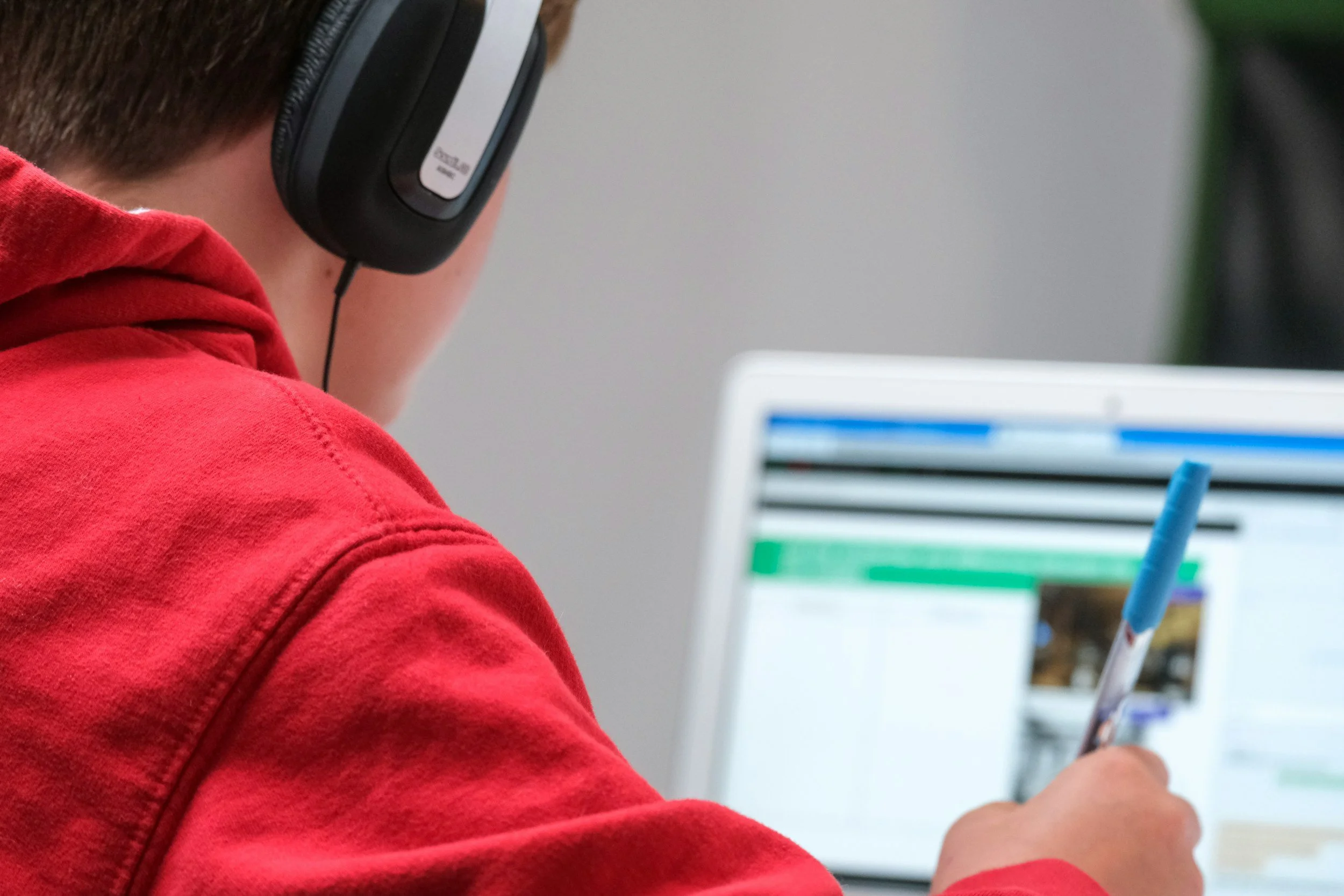 A person wearing a red hoodie and black headphones sitting at a desk, working on a computer with a white monitor, holding a blue marker, and looking at a screen displaying a webpage or document.