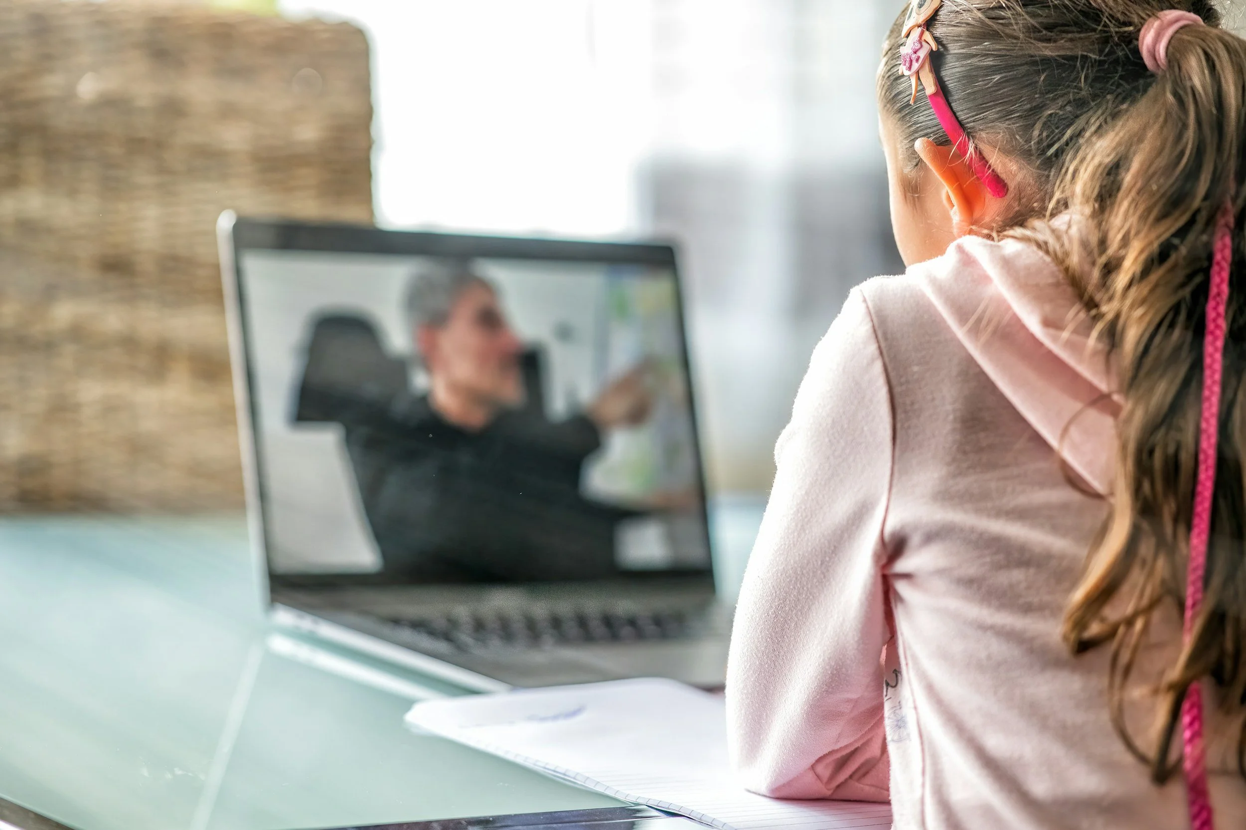 A young girl with glasses sitting at a desk on a video call with an older woman on a laptop, with the woman slightly blurred in the background.