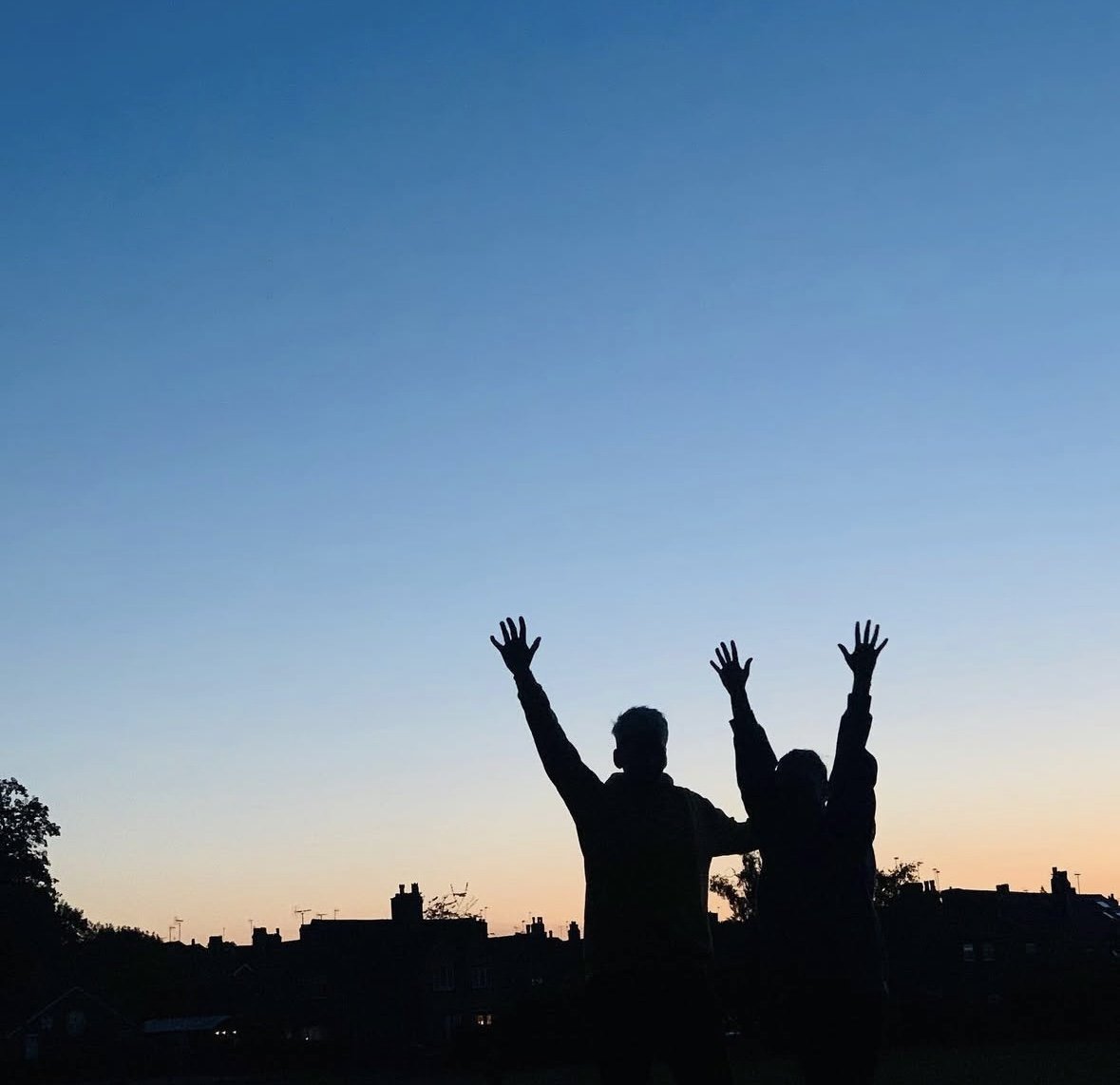 Silhouettes of two people with arms raised against a sunset sky over a cityscape.