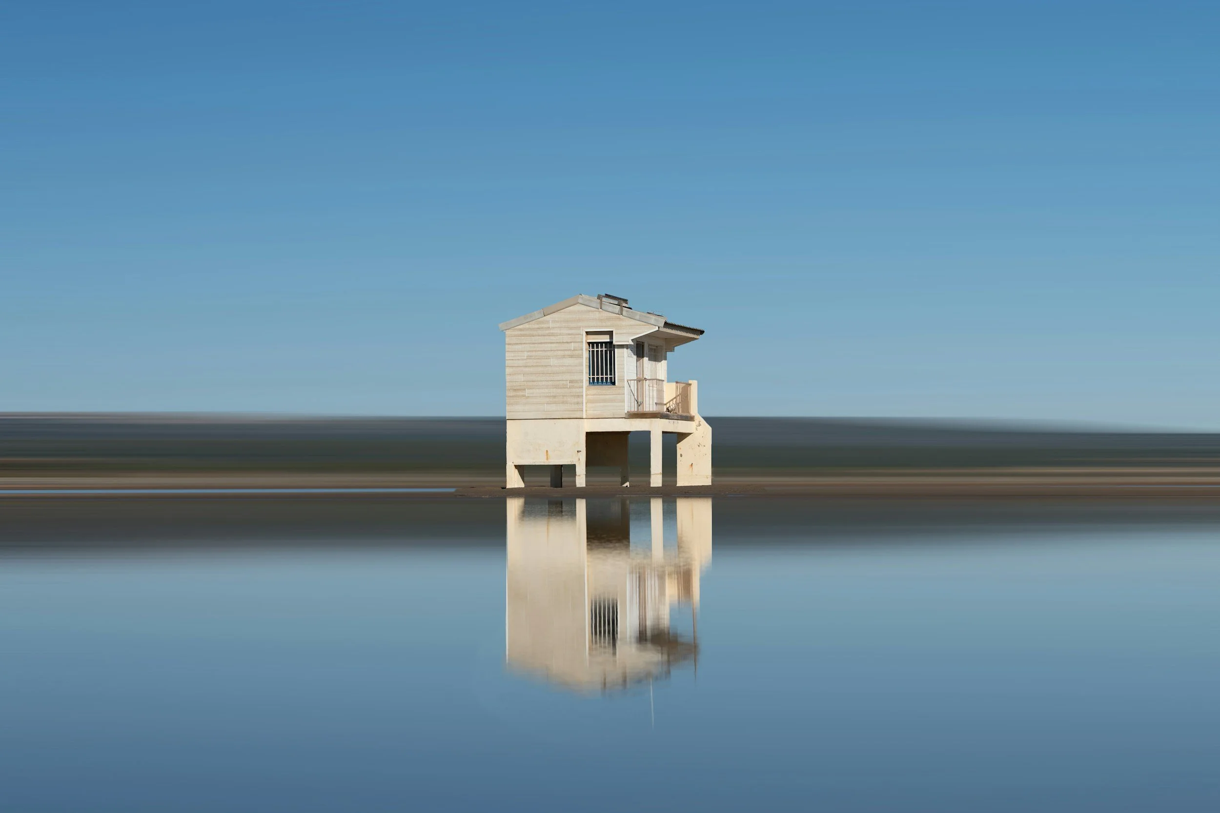 Petit chalet à Gruissan sur pilotis au-dessus de l'eau, avec un reflet dans la surface calme de la mer, sous un ciel bleu.