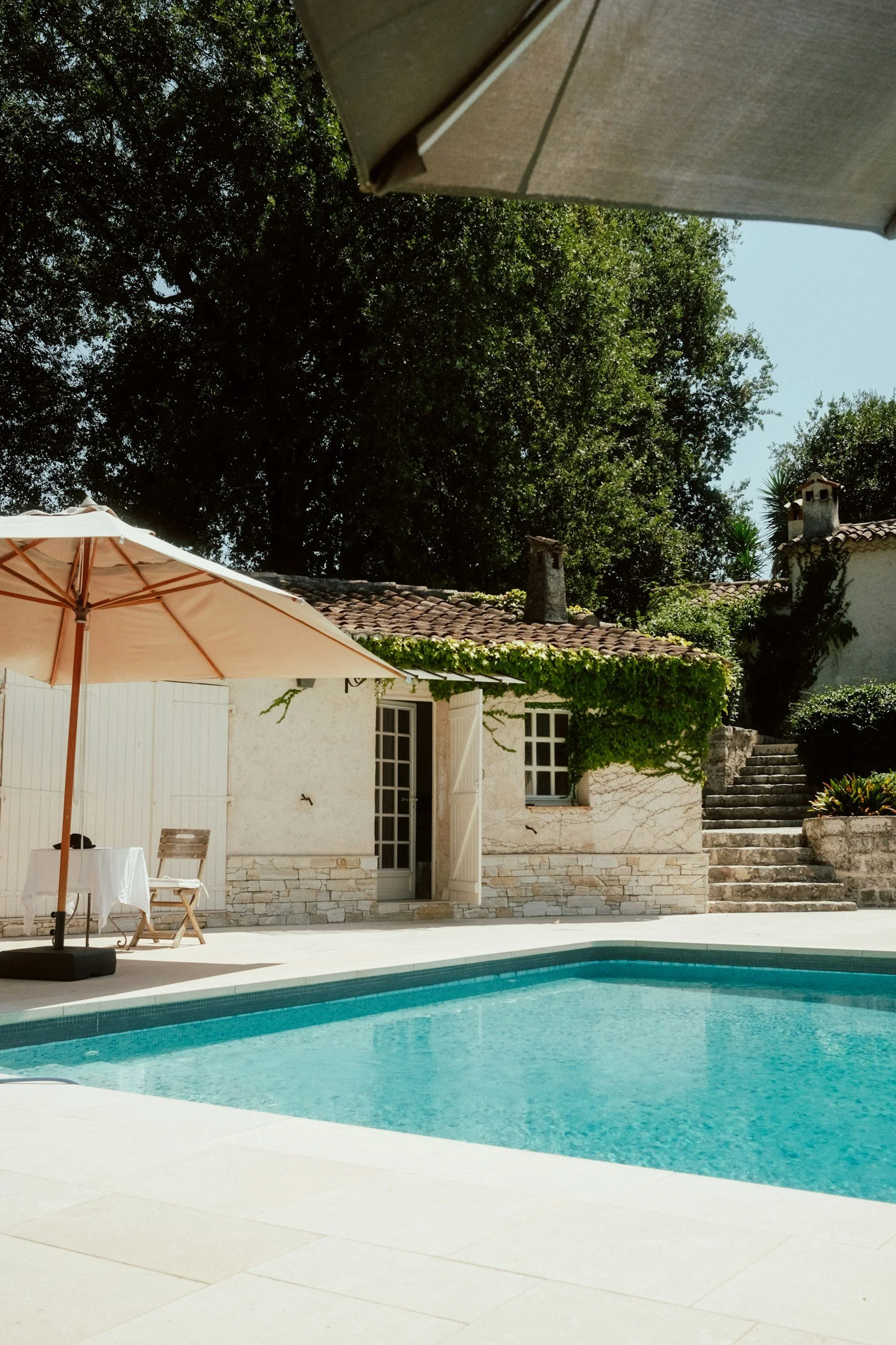 Une piscine, une maison en pierre avec une porte et des fenêtres, un parasol beige, une table avec une nappe blanche, une chaise en bois, et des escaliers en pierre entourés de végétation.