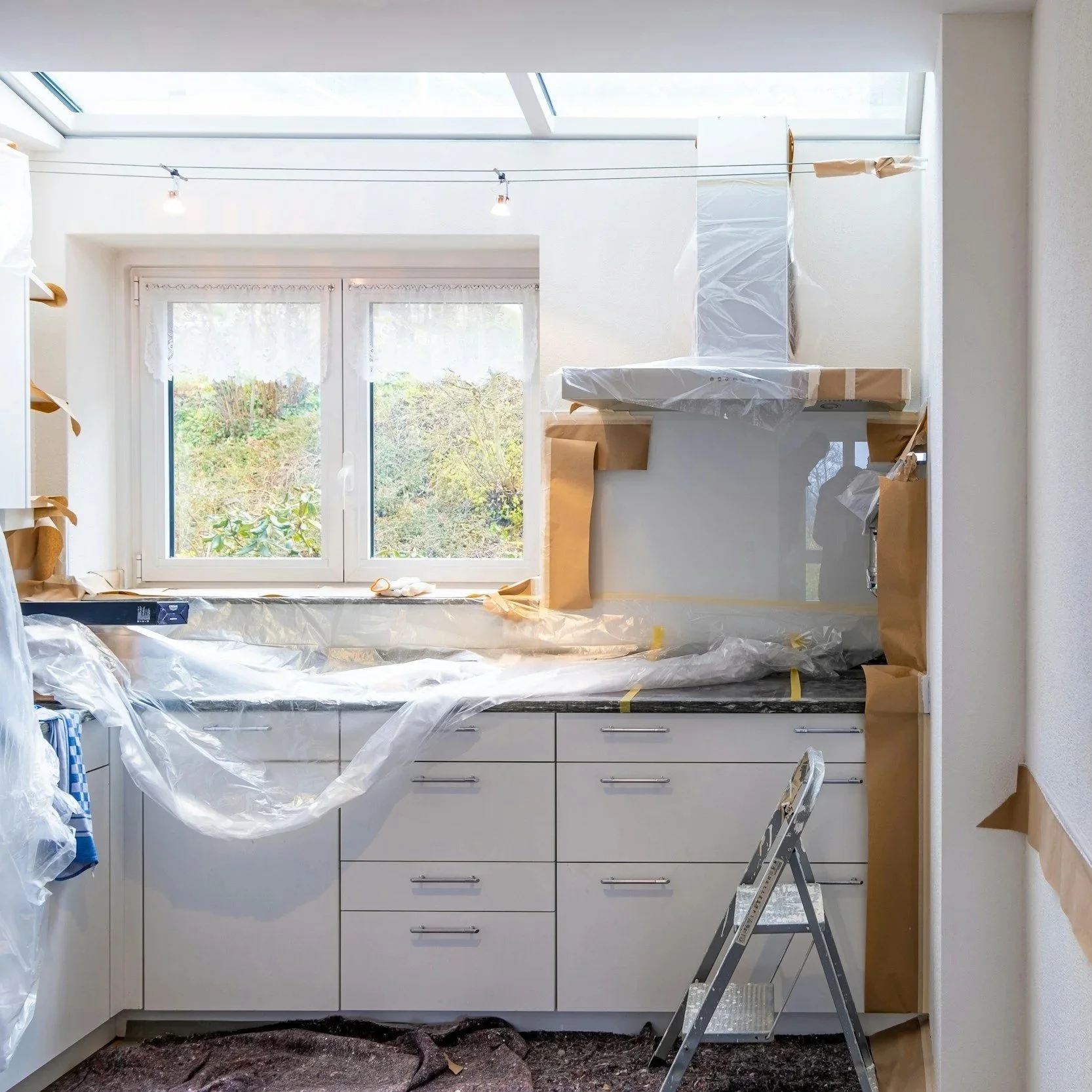Cuisine en rénovation avec des matériaux de protection en plastique sur le plan de travail, une échelle en métal, fenêtre, un mur encadré