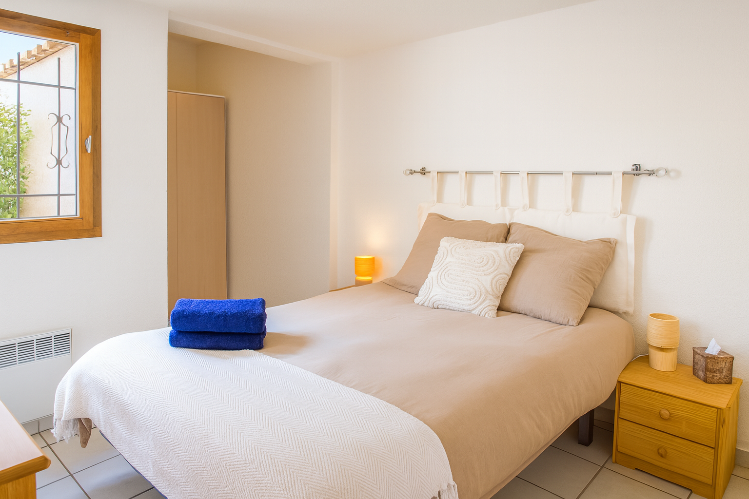 Chambre cosy avec un lit beige, deux luminaires orange, une table de nuit en bois, un oreiller blanc avec motif, et une fenêtre laissant entrer de la lumière naturelle.