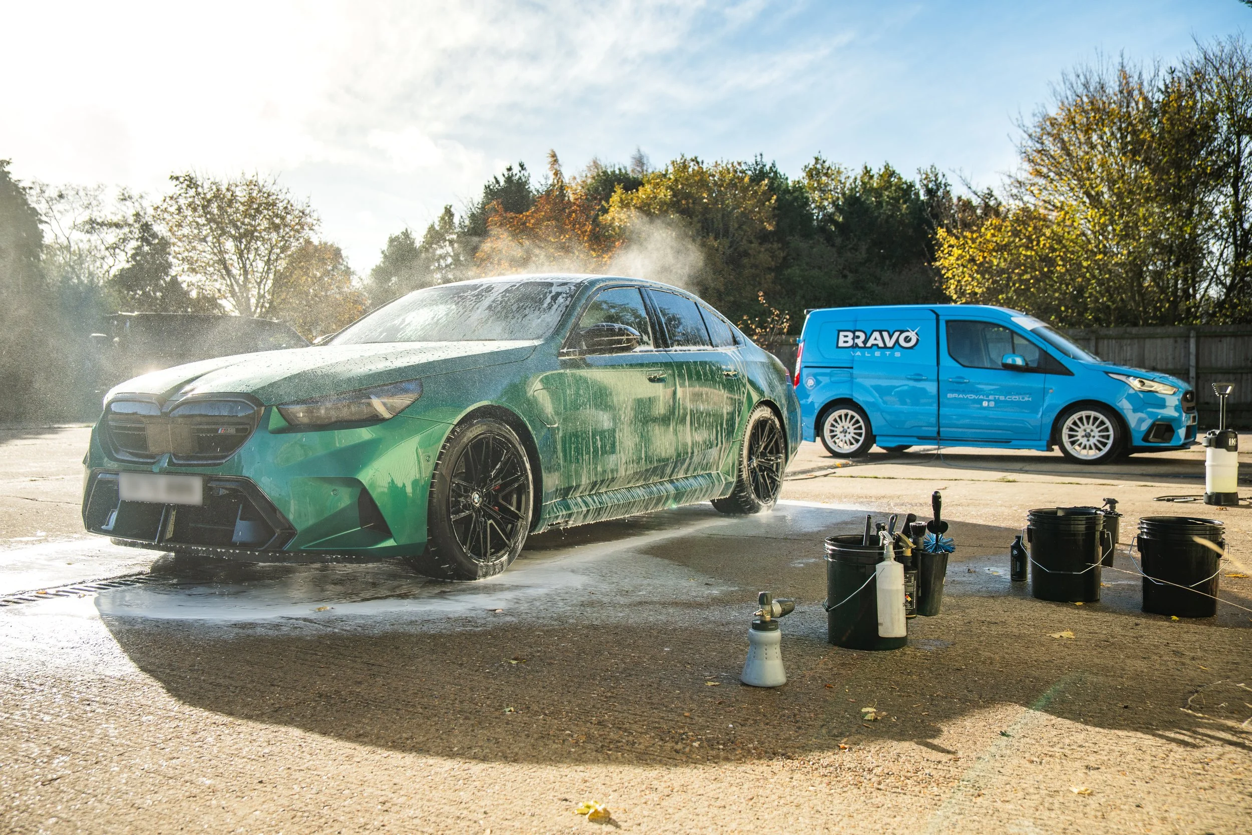 Person washing a gray sports car with a hose on a sunny day in an outdoor car wash.