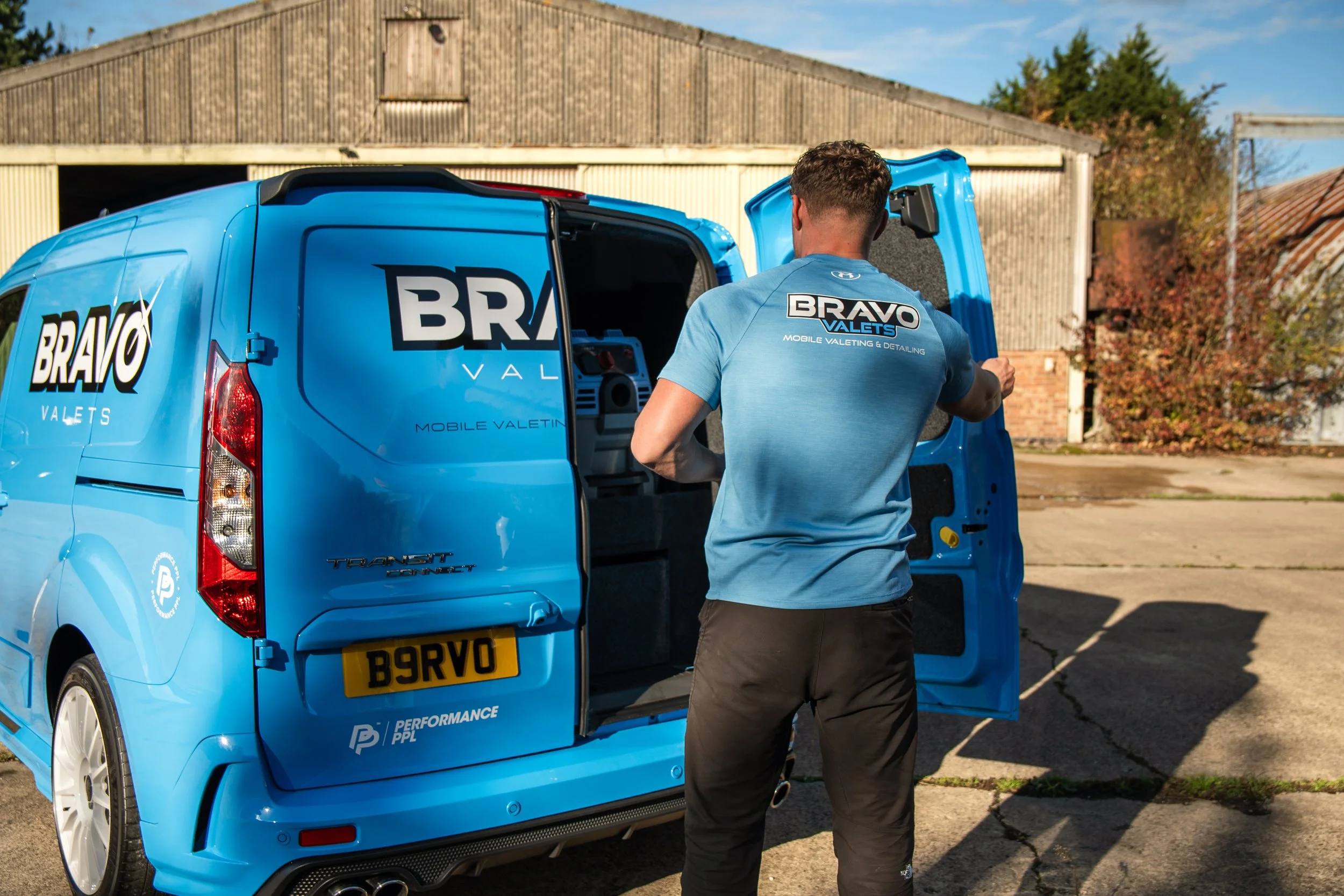 A man in a blue t-shirt with 'BRAVO VALETS' logo on the back is standing next to a blue van with the same logo, opening the rear doors. The van has a yellow license plate with B9RVO and is parked outdoors near a warehouse.