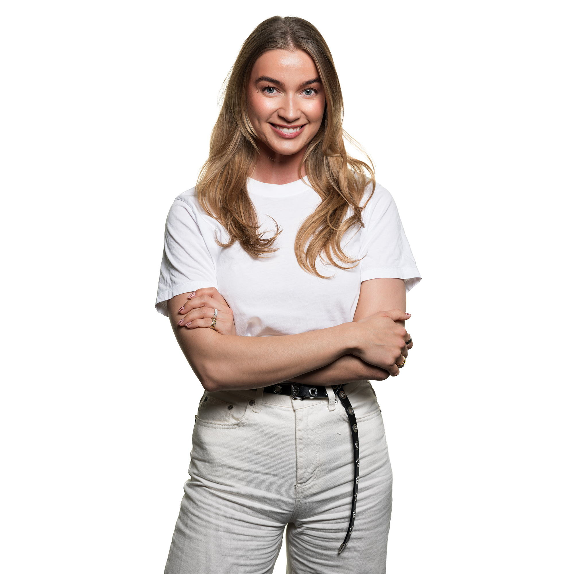 A woman with long, wavy blonde hair wearing a white t-shirt and light-colored pants, smiling and crossing her arms, against a plain white background.