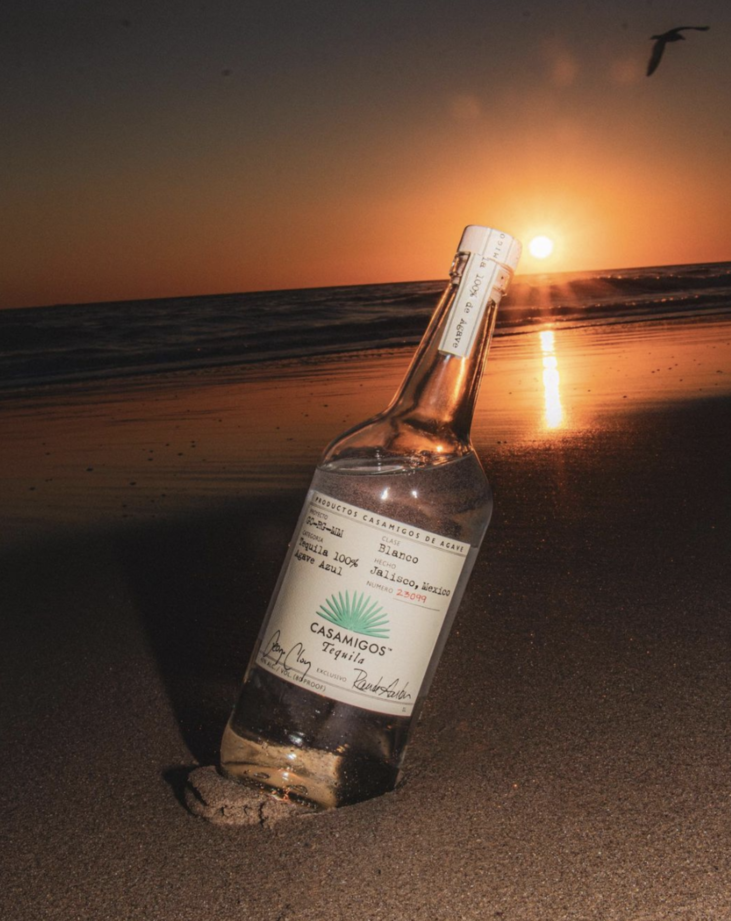 A bottle of Casamigos Blanco Tequila nestled upright in the wet sand of a beach, with the sun setting over the ocean horizon in the background and casting a warm golden glow across the water.