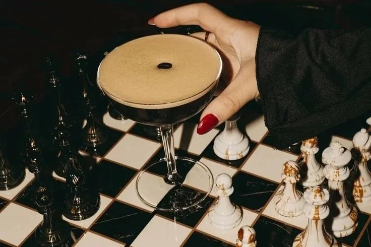 Hand with red nails holding an espresso martini over a marble chessboard, symbolizing precision, balance, and strategy in decision-making.