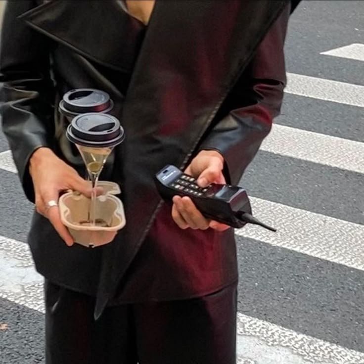 Street-style shot of a person in a black leather outfit holding coffee cups and a retro phone — blending modern hustle with nostalgic aesthetics.