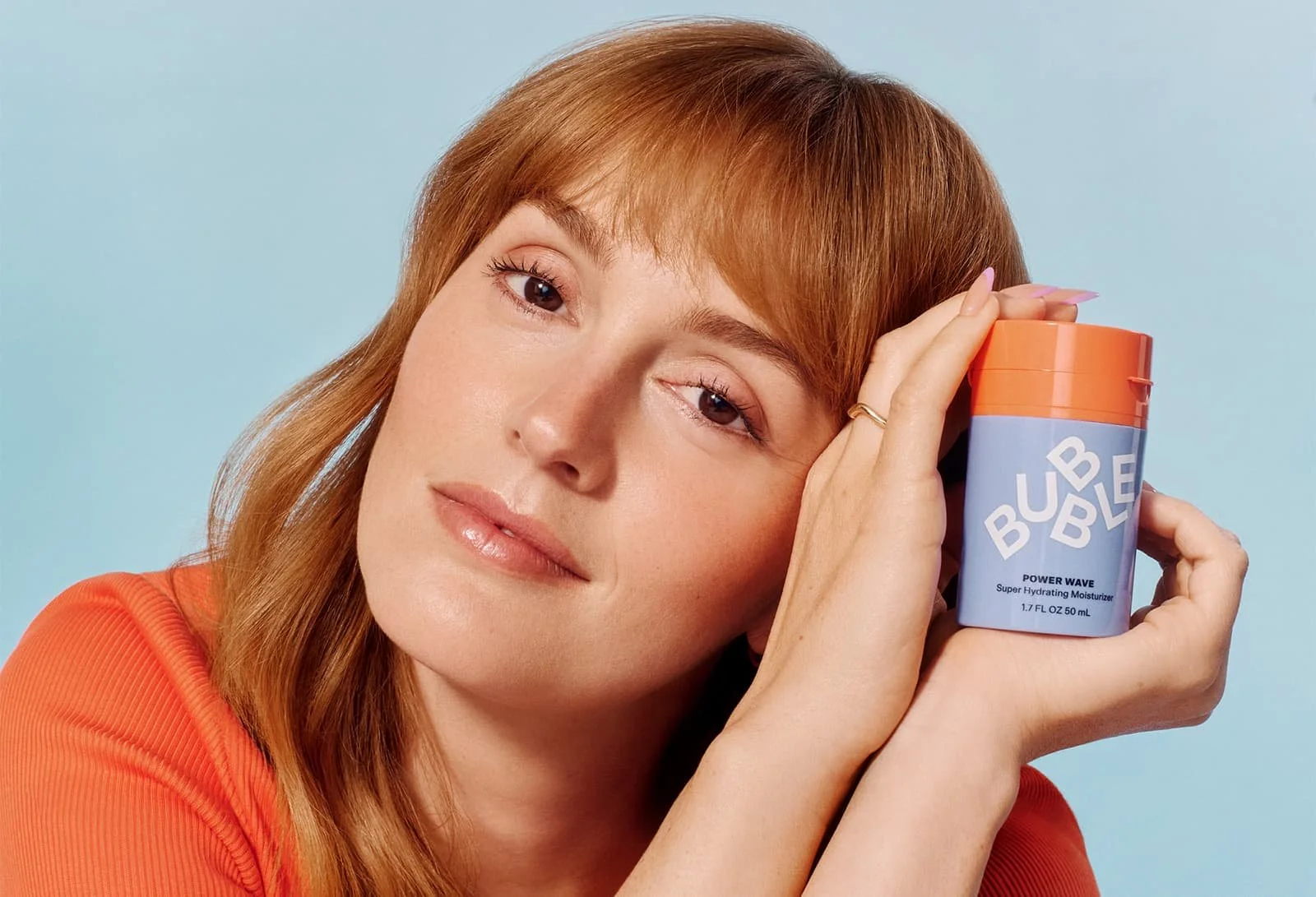 Close-up portrait of a woman holding a pastel-colored Bubble skincare product against a soft blue background, featuring glowy minimal makeup and playful Gen-Z aesthetic.